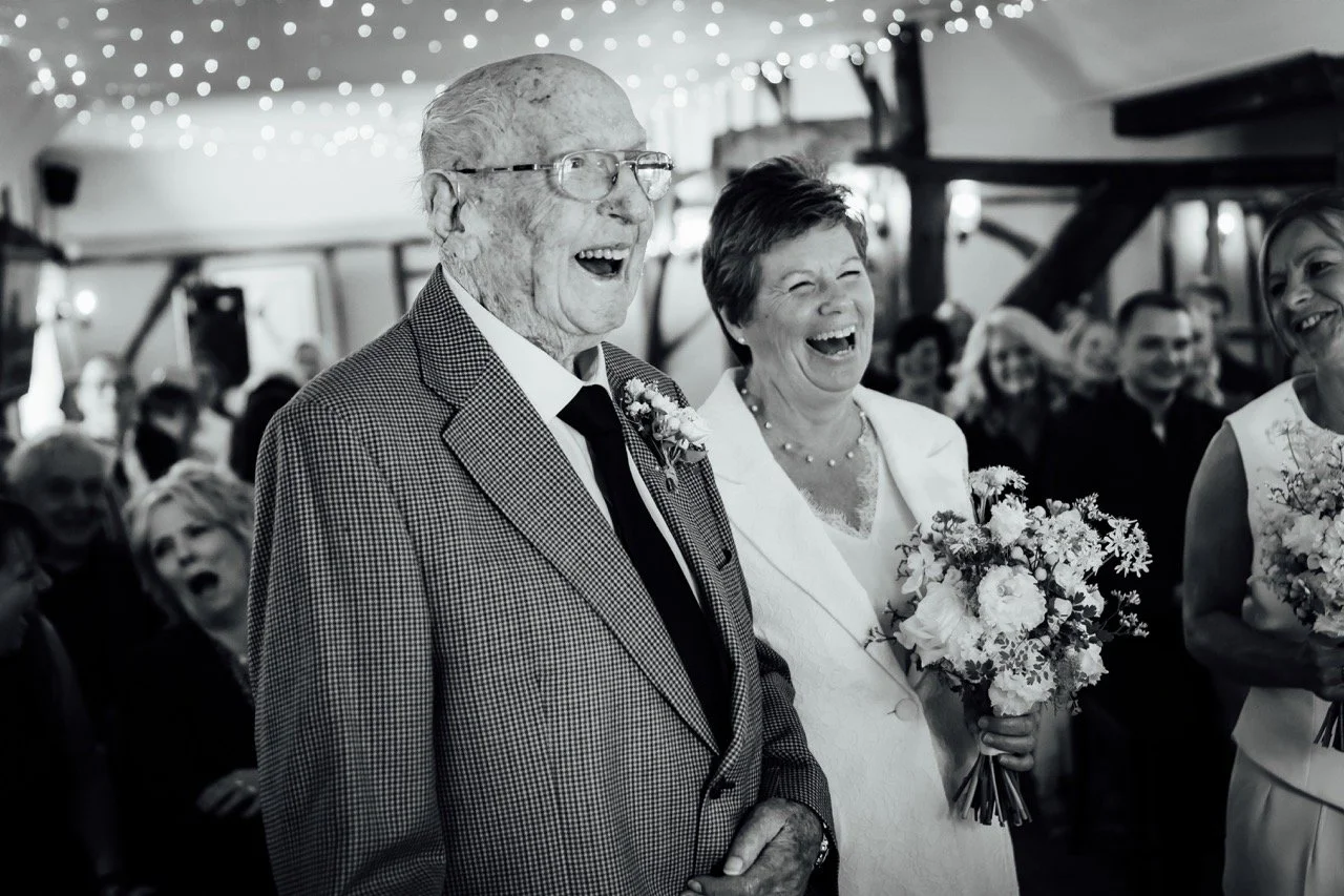 An elderly man and woman, both smiling and laughing, standing at a wedding ceremony. The man is wearing glasses, a checkered blazer, and a boutonniere, while the woman is holding a bouquet of flowers and wearing a white outfit. Other guests in the ba