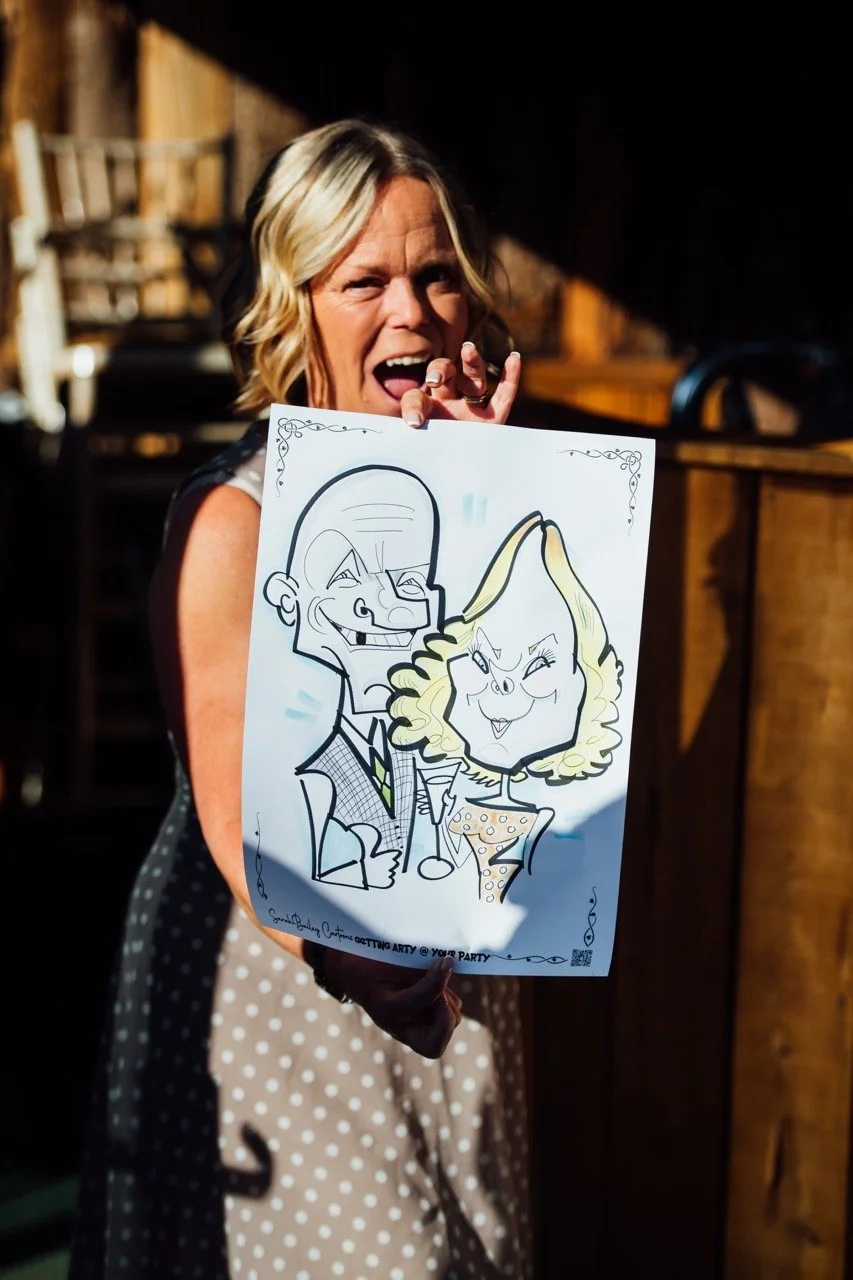 Woman holding a caricature drawing of herself and a man, smiling and pointing at the drawing.