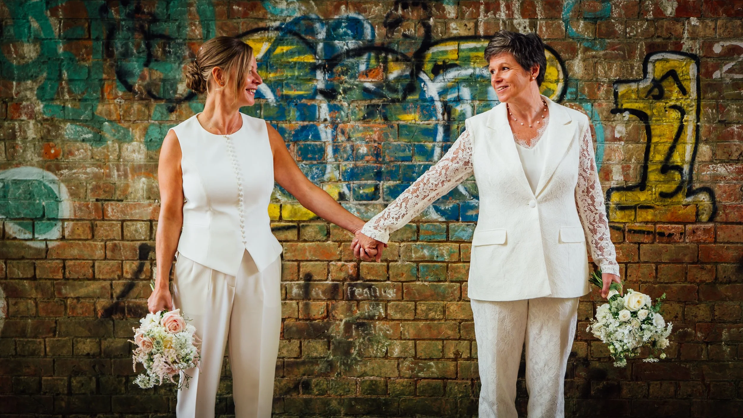 Two women dressed in white, holding bouquets of flowers, holding hands and smiling at each other in front of a graffiti-covered brick wall.