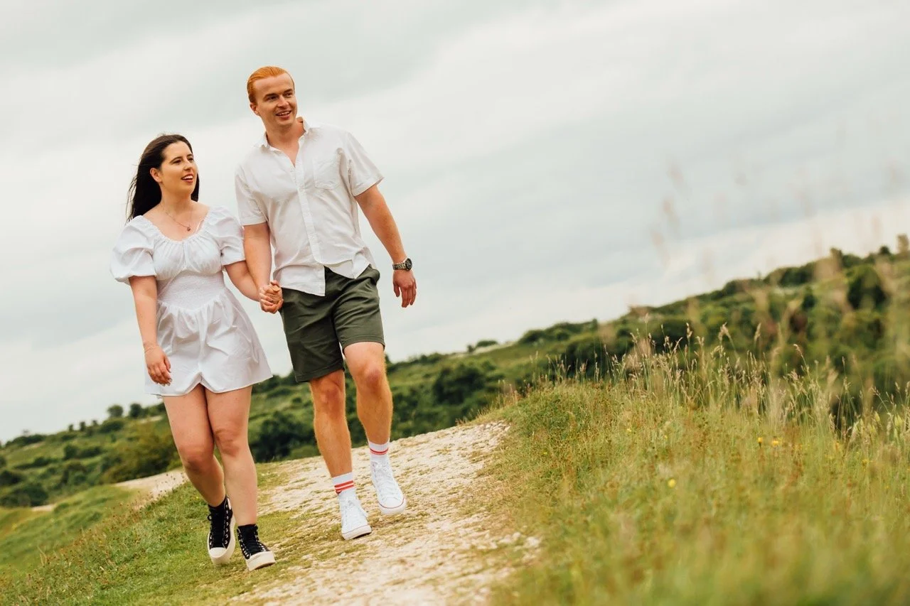 A young couple walking hand-in-hand on a dirt trail in a grassy field with rolling hills and cloudy sky in the background.