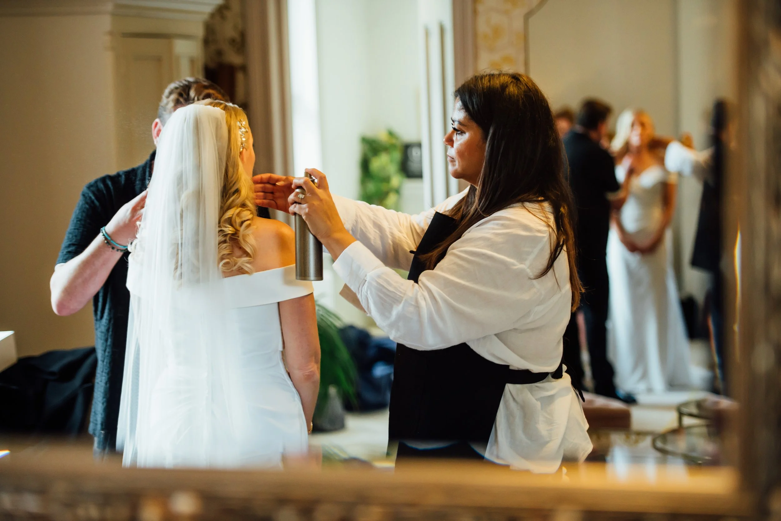 A woman with long dark hair preparing a bride for a wedding, applying makeup or fixing her hair, while the bride is dressed in a white off-the-shoulder gown with a veil. Several other women are in the background, also dressed in white wedding dresses