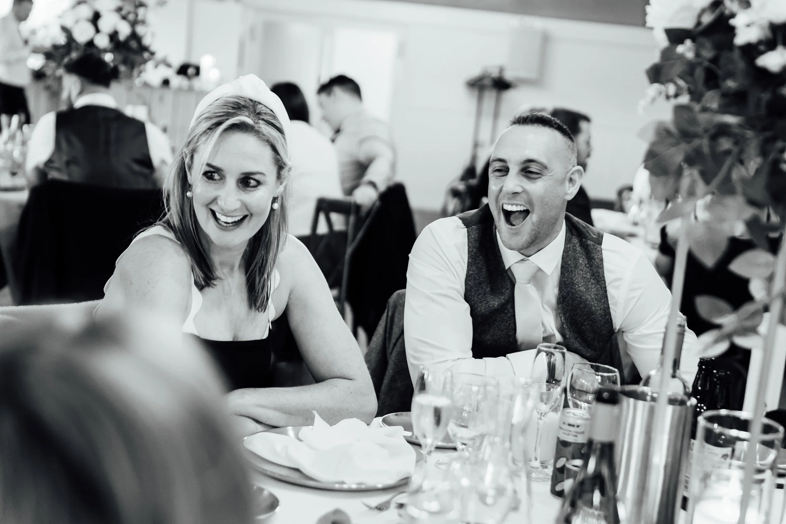 A black and white photo of a woman and a man sitting and smiling at a table at a social gathering or celebration, with other people in the background.
