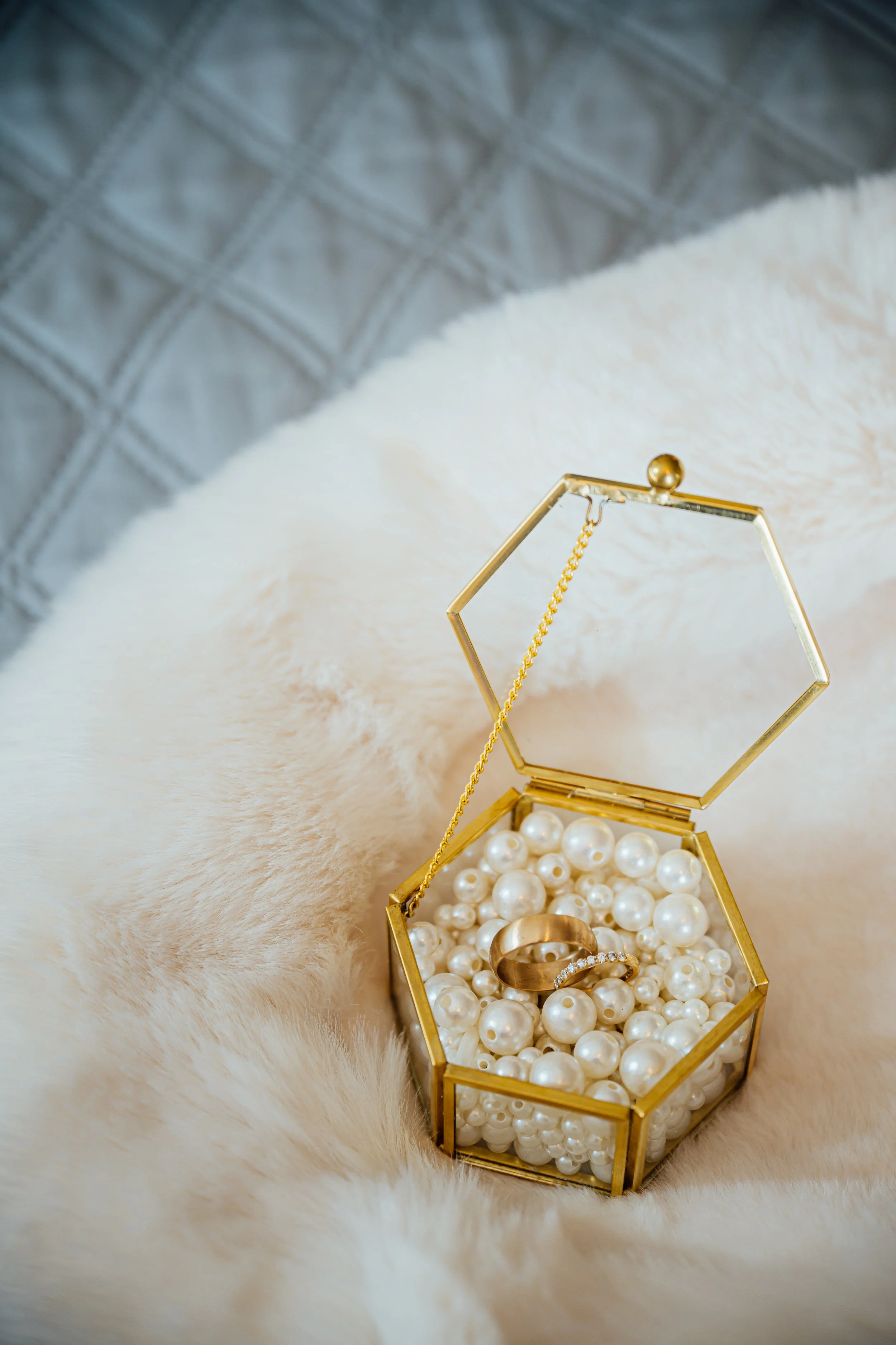 A gold jewelry box filled with pearls and a gold ring with diamonds, placed on a soft, cream-colored fur surface.