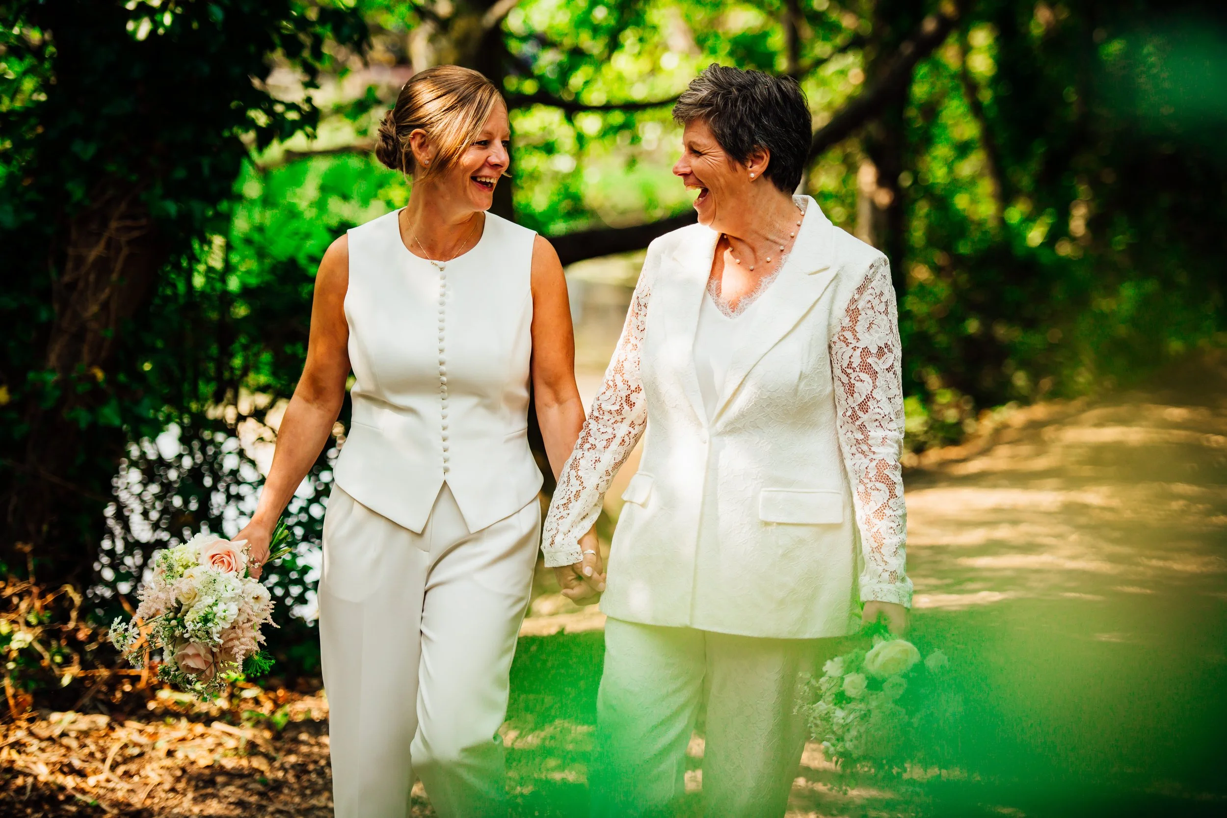 Two women in white outfits holding hands and smiling at each other outside in a green, leafy park.