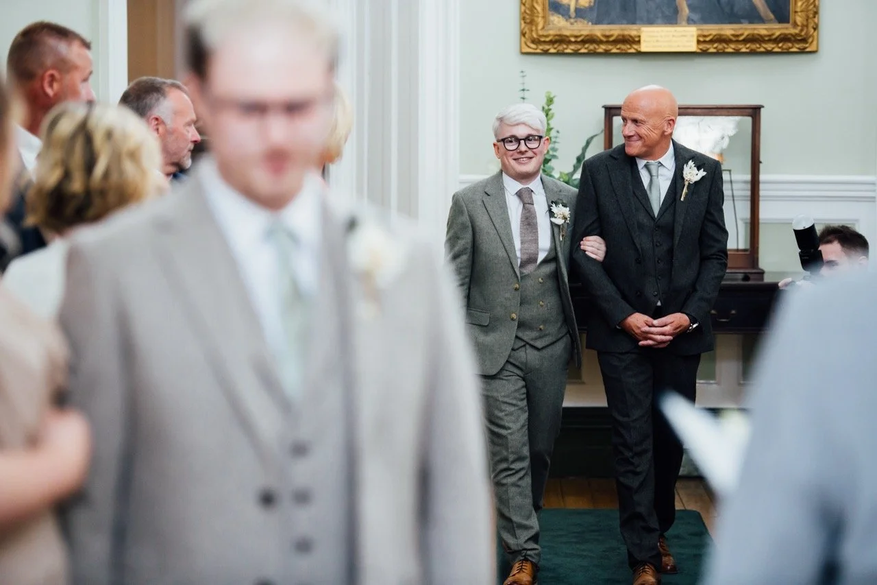 A wedding ceremony with a groom and his father walking down the aisle, holding arms, in a decorated room with guests seated on each side.