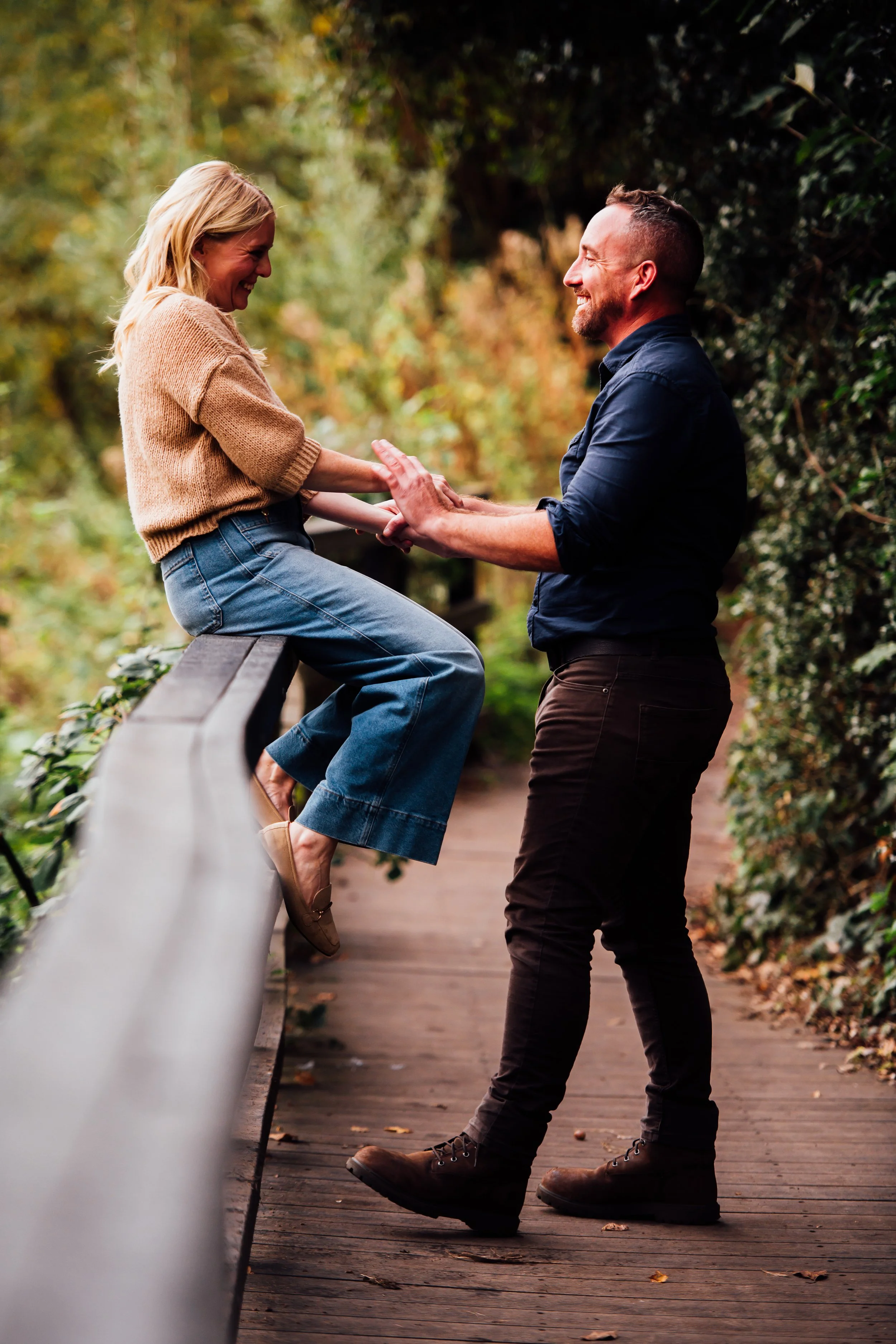 A man and woman smiling and holding hands, with the woman sitting on a wooden railing and the man standing on the ground, in an outdoor park setting with trees and fallen leaves.