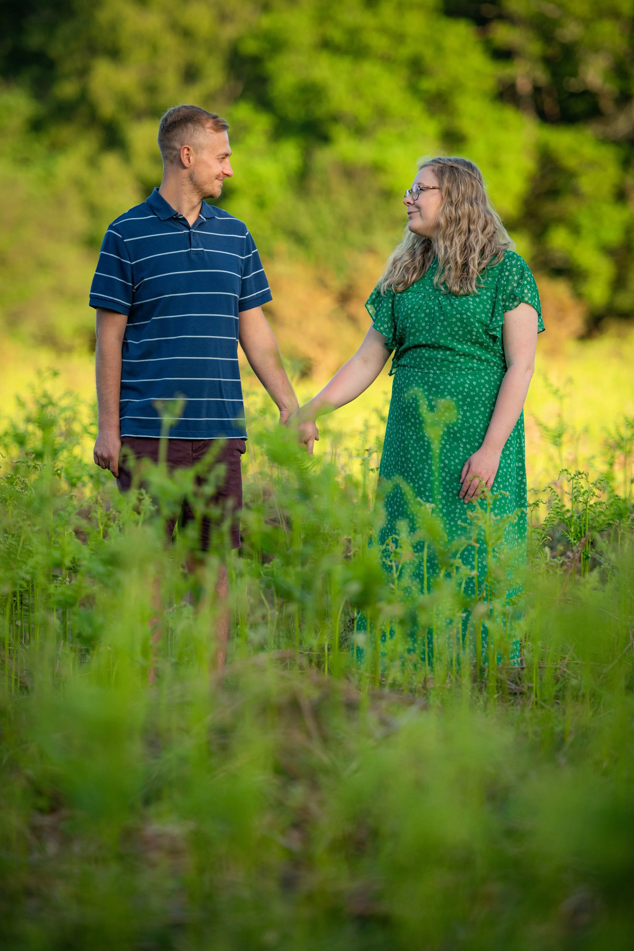A couple holding hands and smiling at each other in a green outdoor setting.