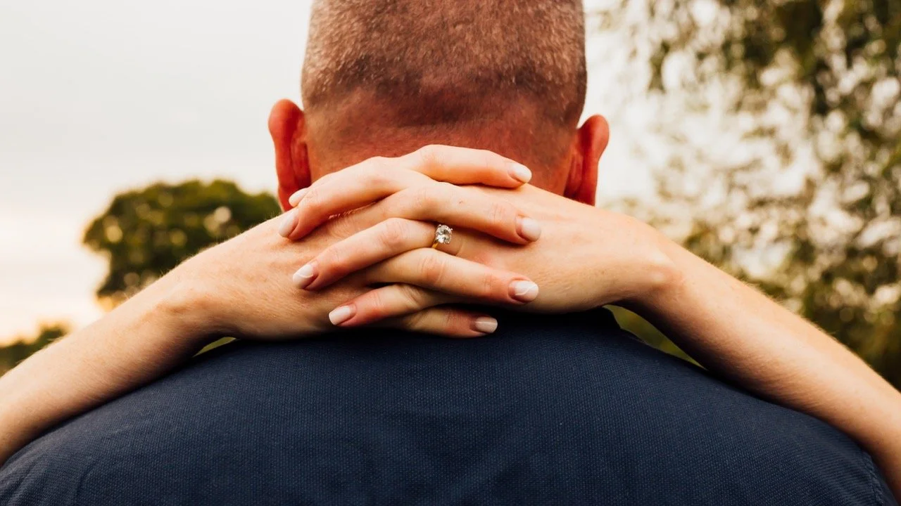 Close-up of a woman with a wedding ring on her finger embracing a man. The woman is hugging the man from behind, with her hands around his neck, and her face is not visible. The background shows trees and a soft, outdoor setting.