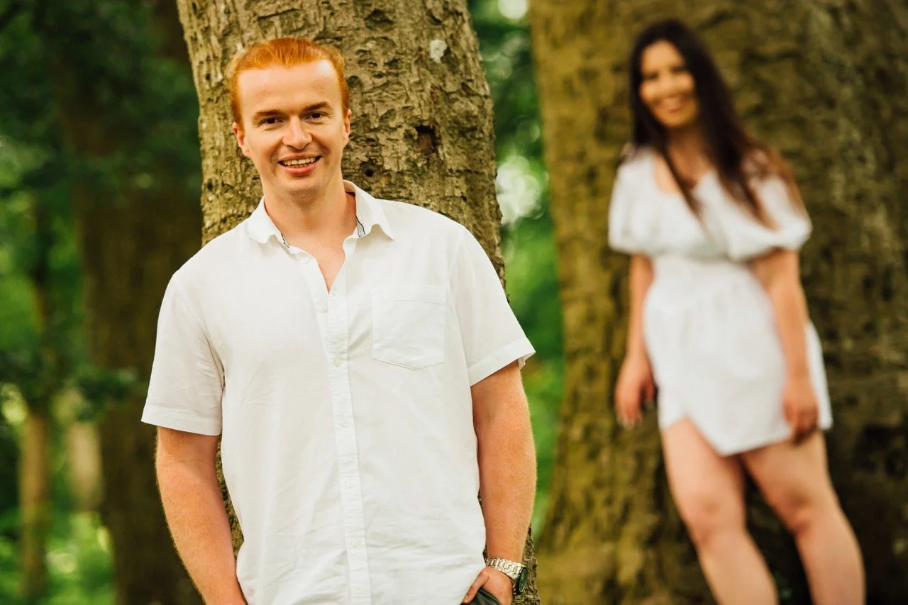 A smiling man with red hair in a white shirt leaning against a tree, with a blurred woman in a white dress in the background also leaning against a tree in a forest setting.