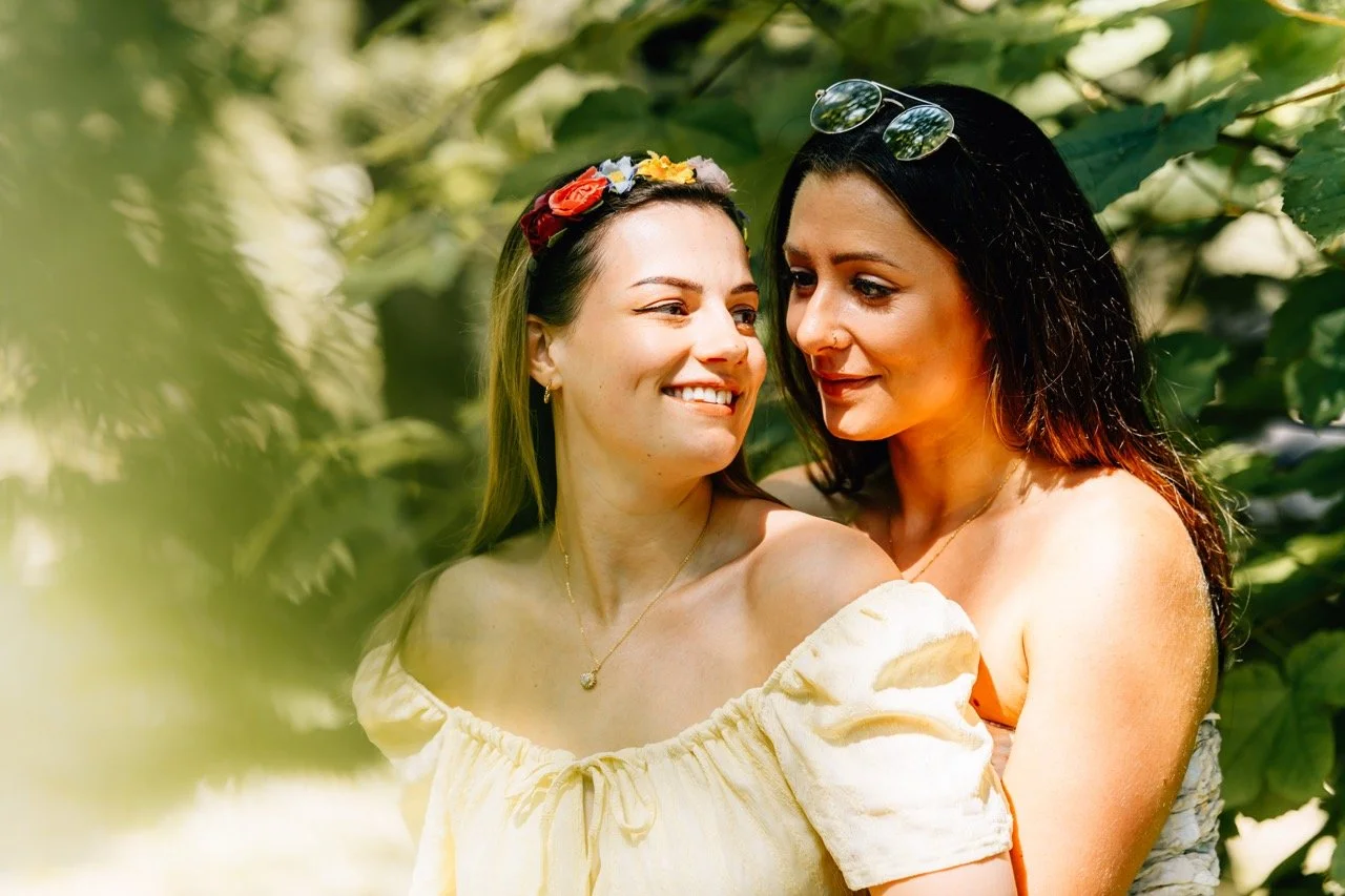 Two women standing close together in a lush green outdoor setting, smiling and looking at each other, with one wearing sunglasses on her head and the other wearing a floral headband.