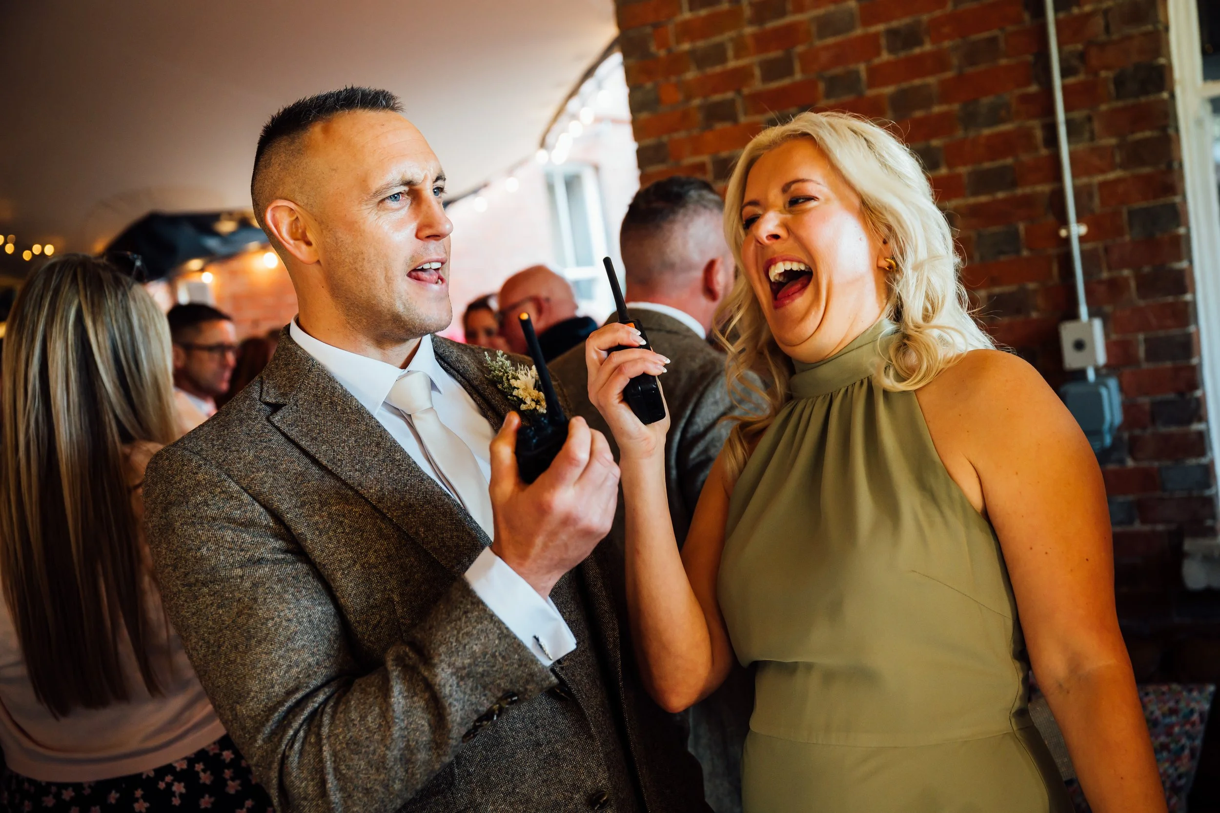 A man and woman laughing and talking at a social event, with other people in the background. The man is holding a walkie-talkie and is dressed in a suit, while the woman is wearing a sleeveless green dress and has blonde hair.