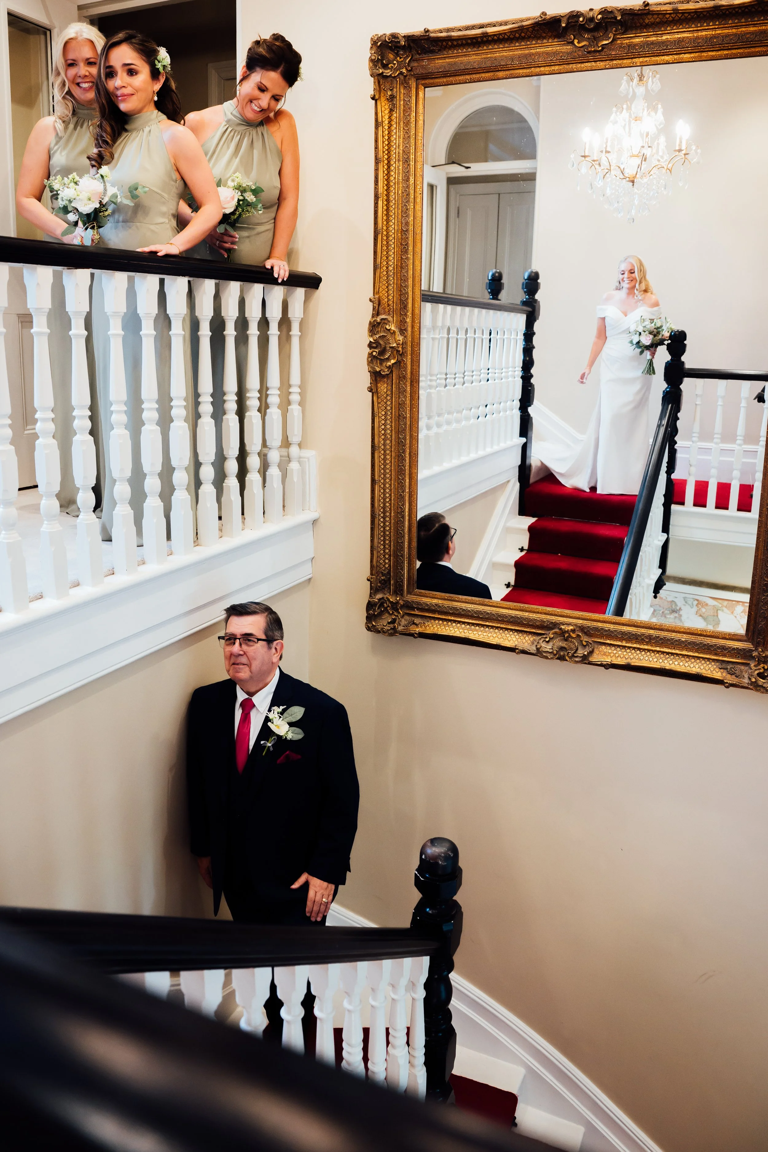 A bride walking down the staircase in a white wedding dress, with bridesmaids looking on from the upper level, and a man in a tuxedo standing below.