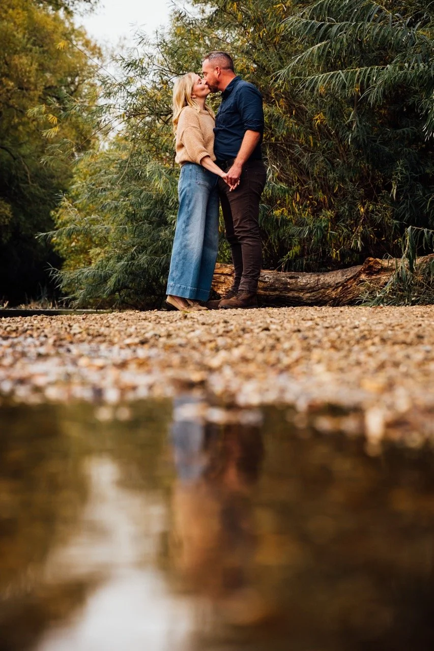A couple kissing while holding hands outdoors among trees, with a creek in the foreground reflecting their image.