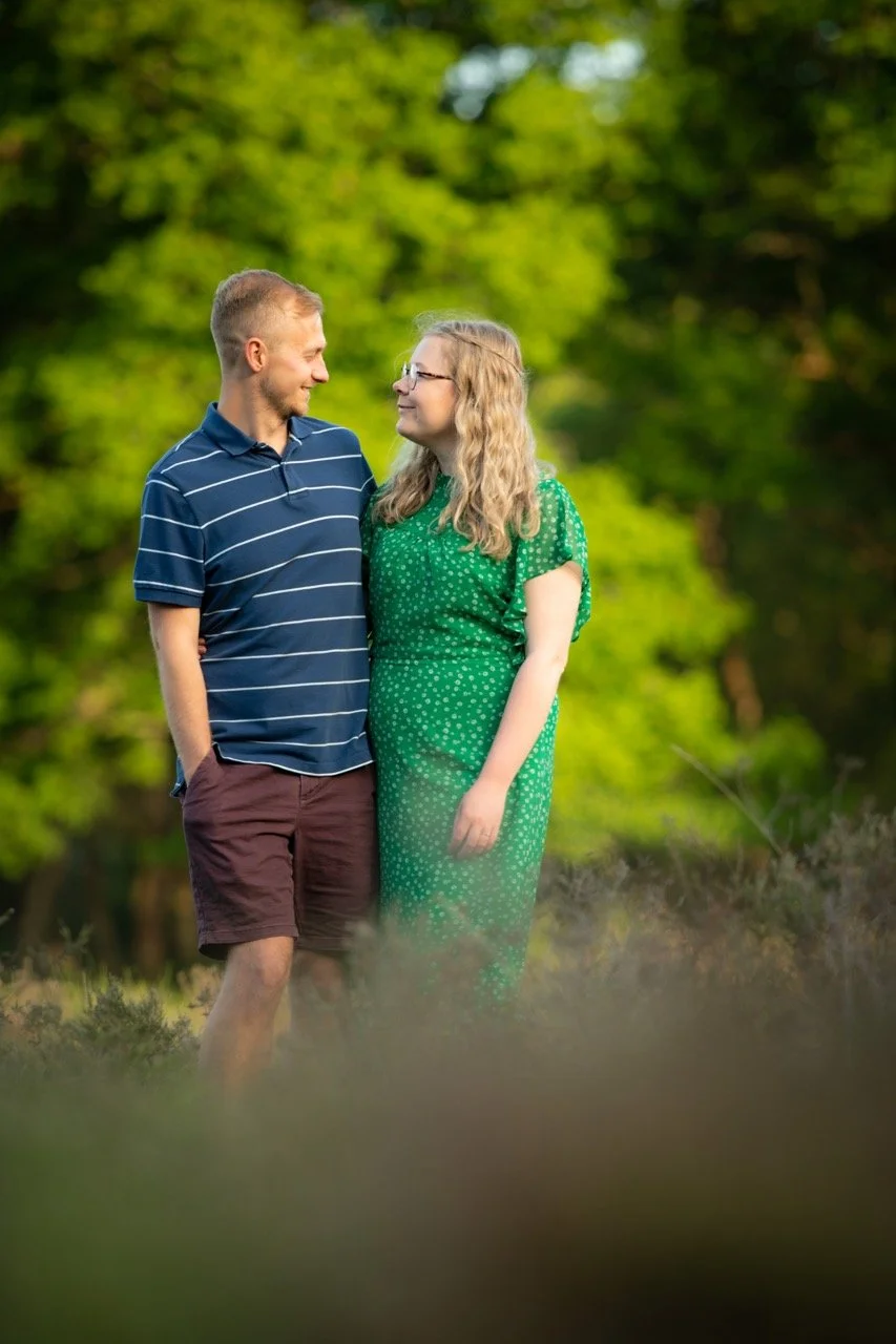 A young man and woman standing close together outdoors, looking at each other, surrounded by greenery.