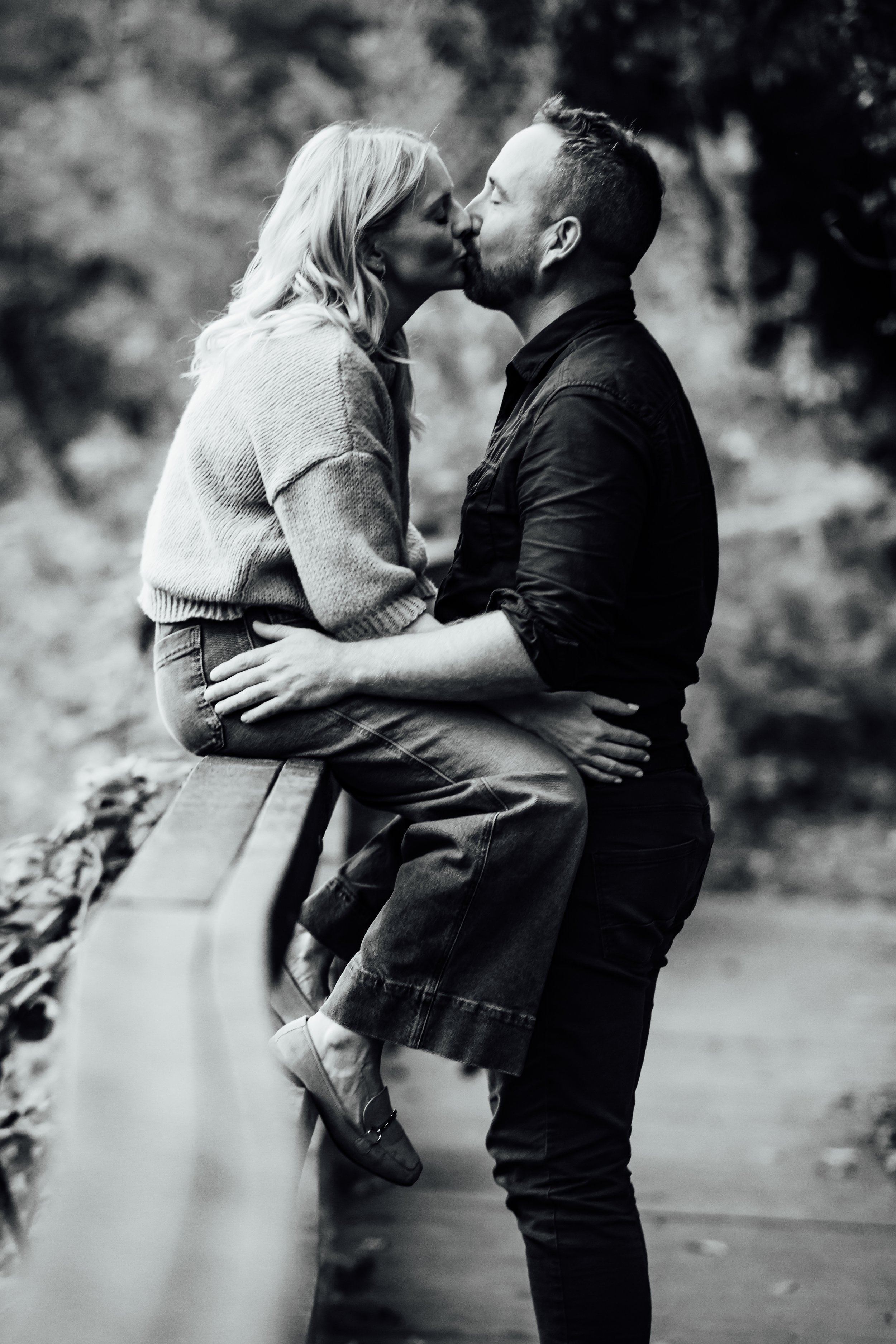 A black and white photo of a couple kissing, with the woman sitting on a park bench and the man standing, holding her waist, outdoors with blurred trees in the background.
