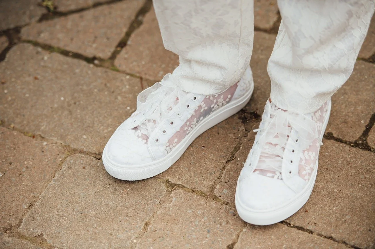 Close-up of white sneakers with lace design and white pants on a brick sidewalk.