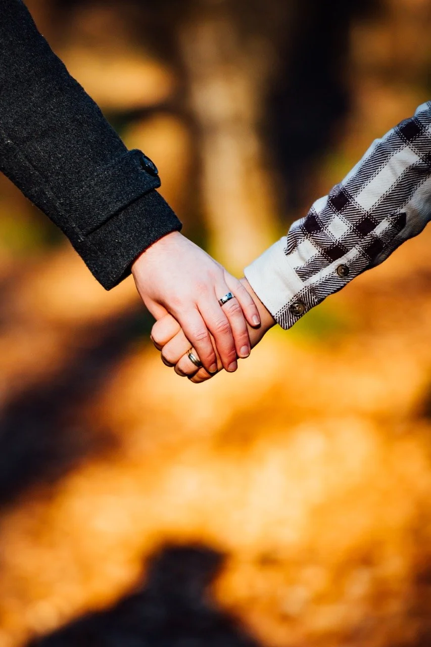 Close-up of two people holding hands, one wearing a black coat and the other wearing a plaid shirt, outdoors with autumn leaves in the background.