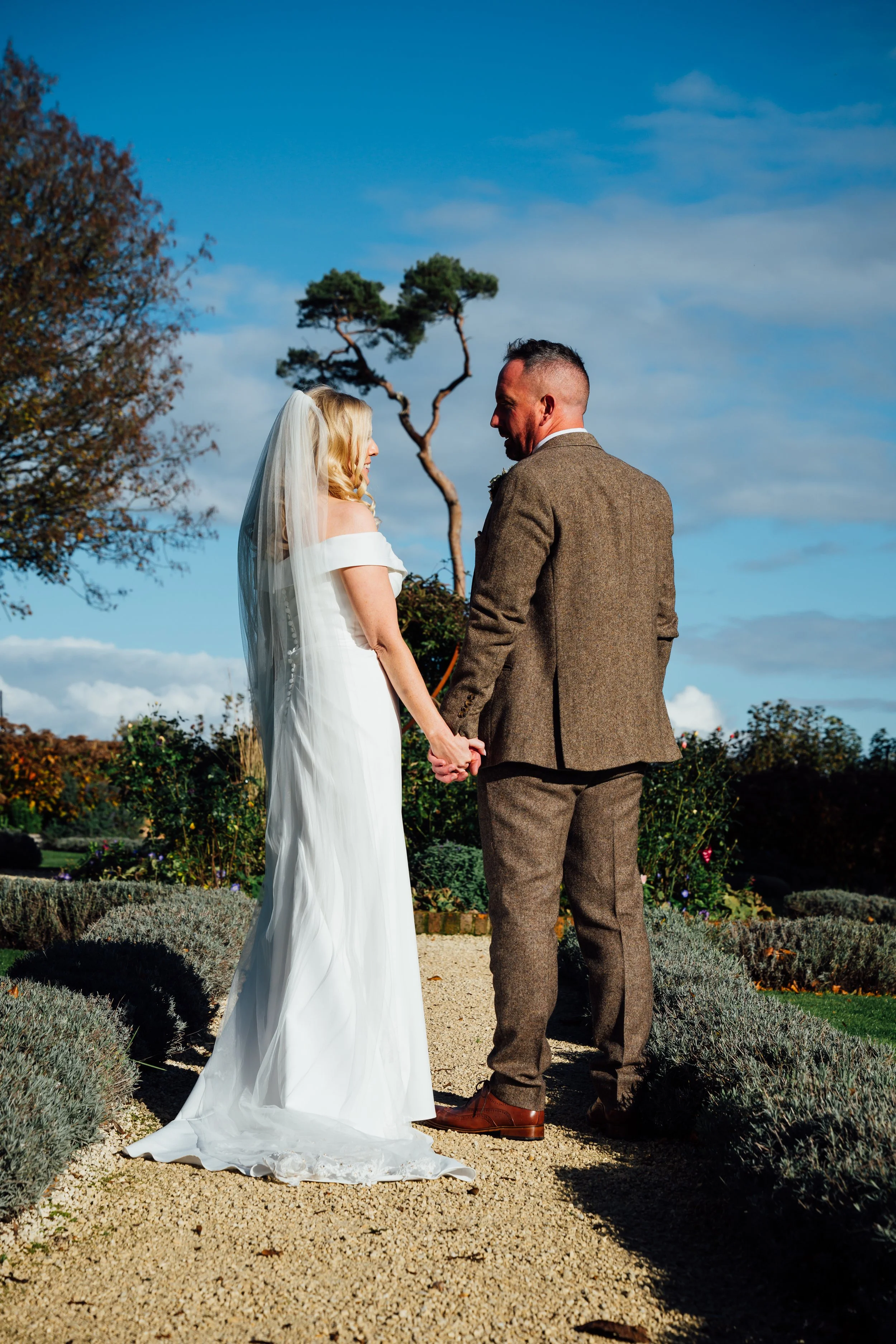 A bride and groom standing hand in hand outdoors on their wedding day, facing each other against a backdrop of trees and a blue sky.