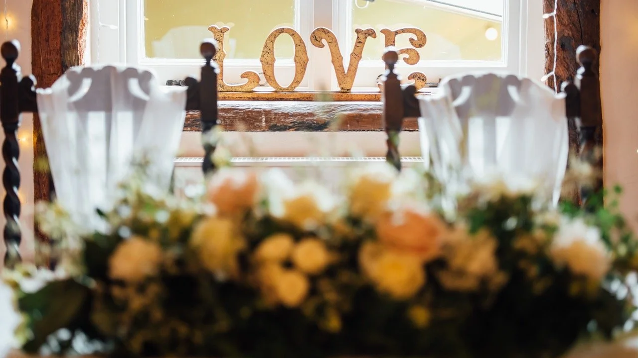 A close-up view of a floral arrangement with white and peach-colored flowers in the foreground, and a window with a wooden frame and white curtains in the background. On the windowsill, there is a decorative sign spelling "LOVE" in large, glittery le