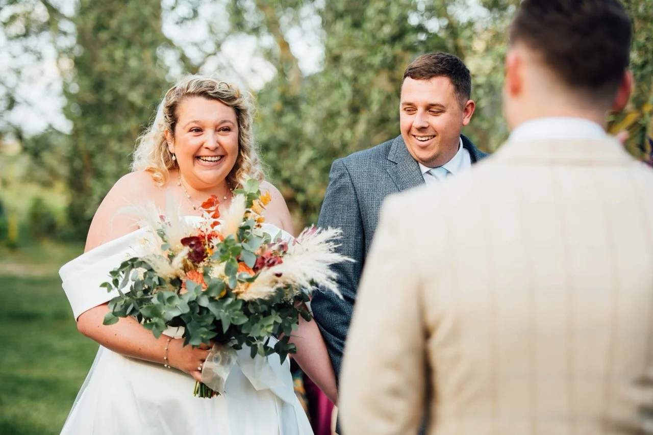 A bride in a white wedding dress holding a bouquet of flowers, smiling at a groom during a wedding ceremony outdoors, with groomsmen and trees in the background.