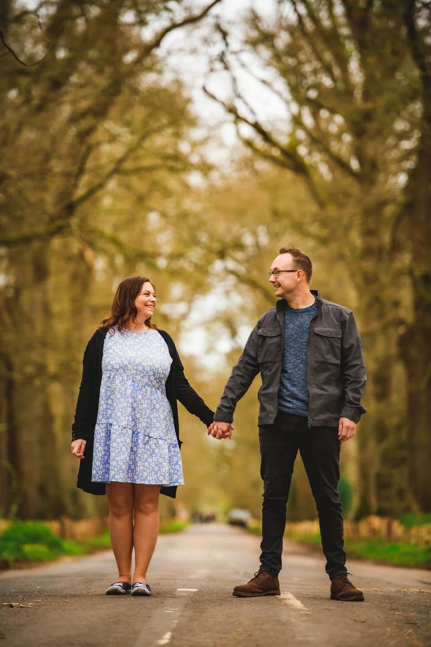 A couple holding hands and smiling at each other on a tree-lined road in autumn.