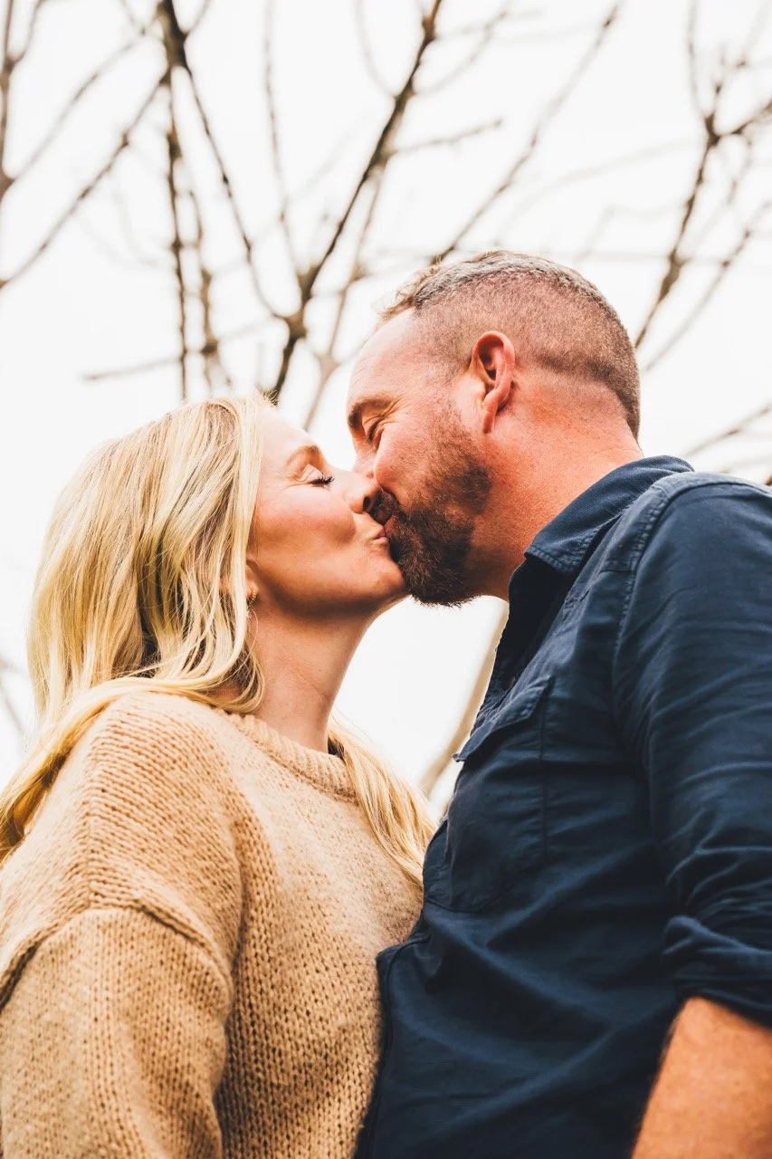 A couple kissing outdoors with bare tree branches behind them.