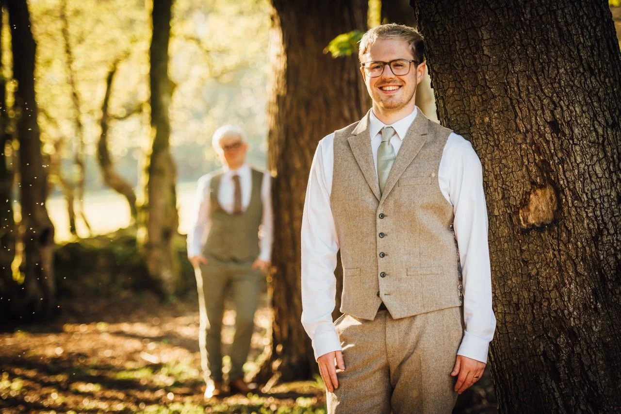 Young man in a tan suit and glasses smiling and leaning against a tree, with an older woman in a vest and tie in the background, in a sunlit forest.