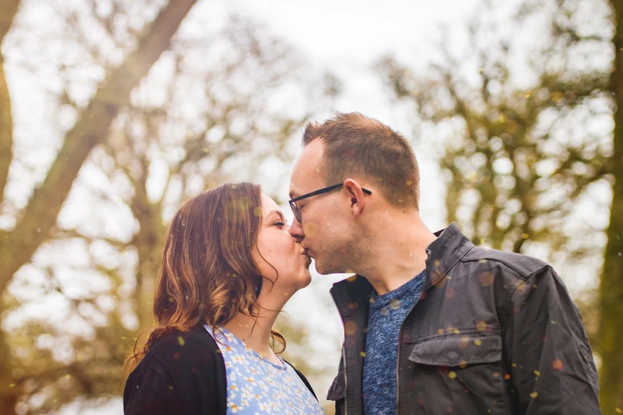 A couple is kissing outdoors with a blurred background of trees and sunlight.