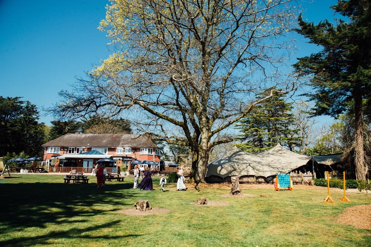 People dressed in vintage clothing standing and walking on a grassy lawn with trees, a large house with a deck, and a tent in the background.