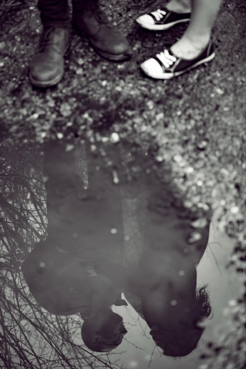 A reflection of a man and a woman in a puddle, with their shoes visible at the top of the image. The ground around them appears to be gravel or dirt.