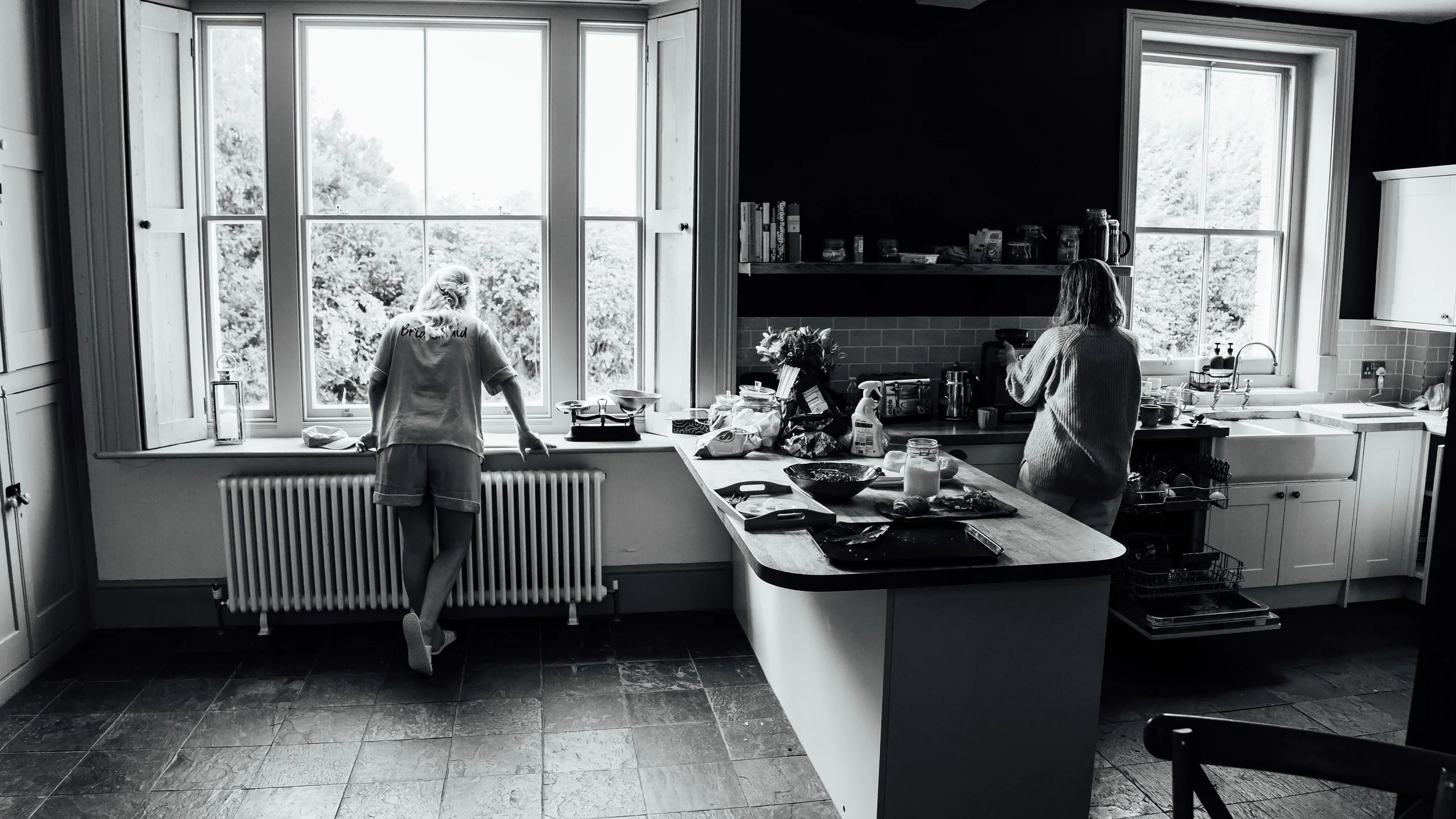 Black and white photo of a kitchen with two women. One is by a large window at the left, looking outside, and the other is at the right, preparing something at the counter. The kitchen has an island with dishes, jars, and cleaning supplies, and an op