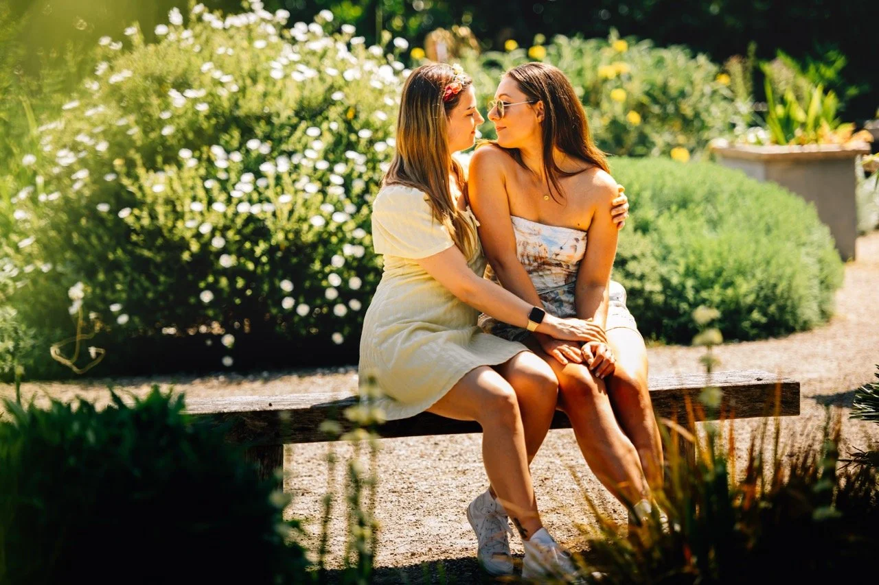 Two women sitting closely on a wooden bench in a garden, sharing an intimate moment, with sunlight, greenery, and flowers in the background.