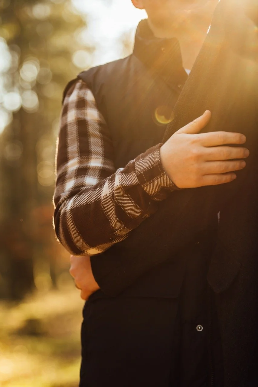 Close-up of a person with one hand over their chest, wearing a plaid shirt and a vest, illuminated by warm sunlight outdoors.
