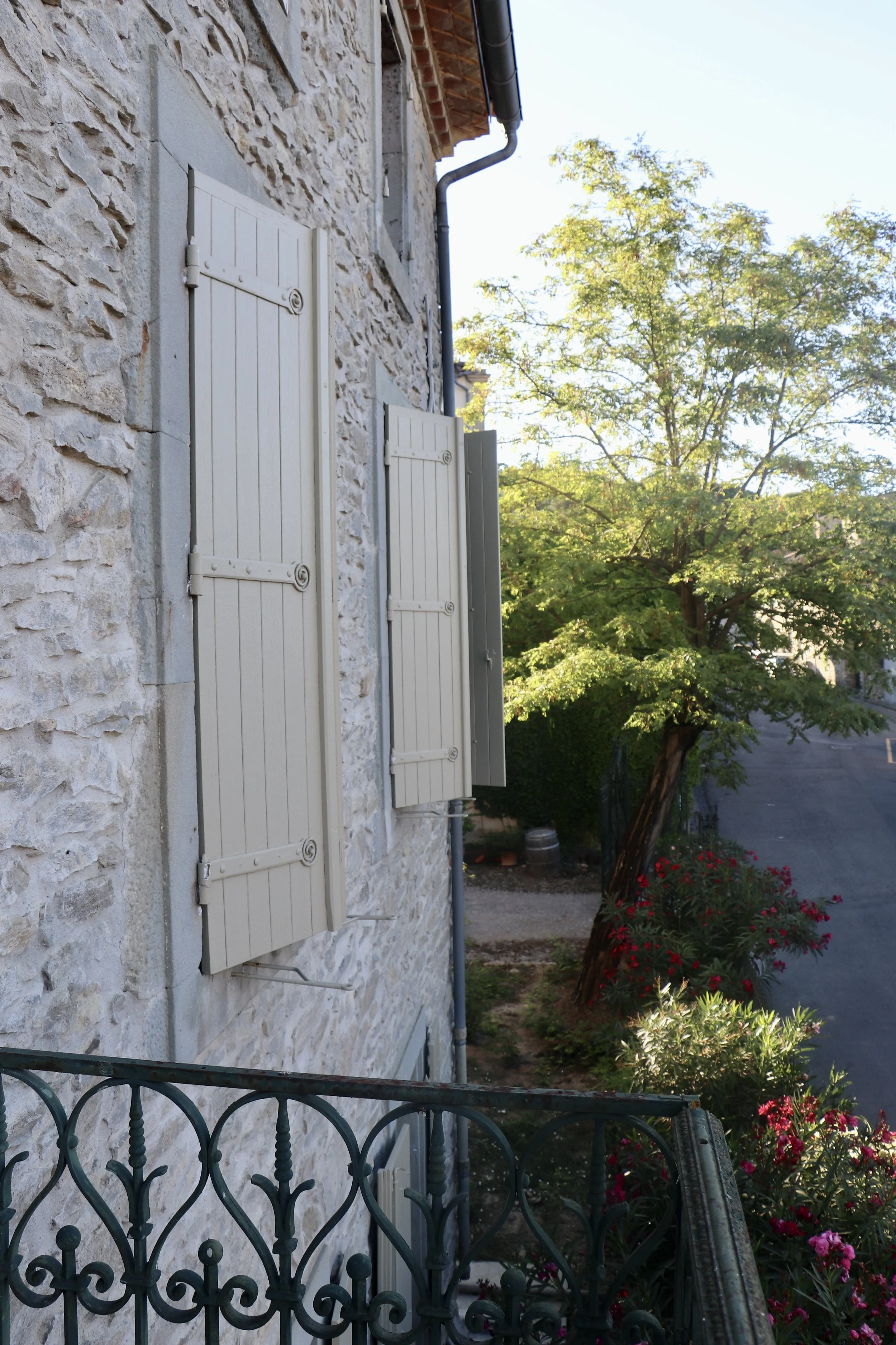 Maison en pierre avec volets en bois blanc, balcon en fer forgé, arbres et fleurs dans un village. Maison de campagne
