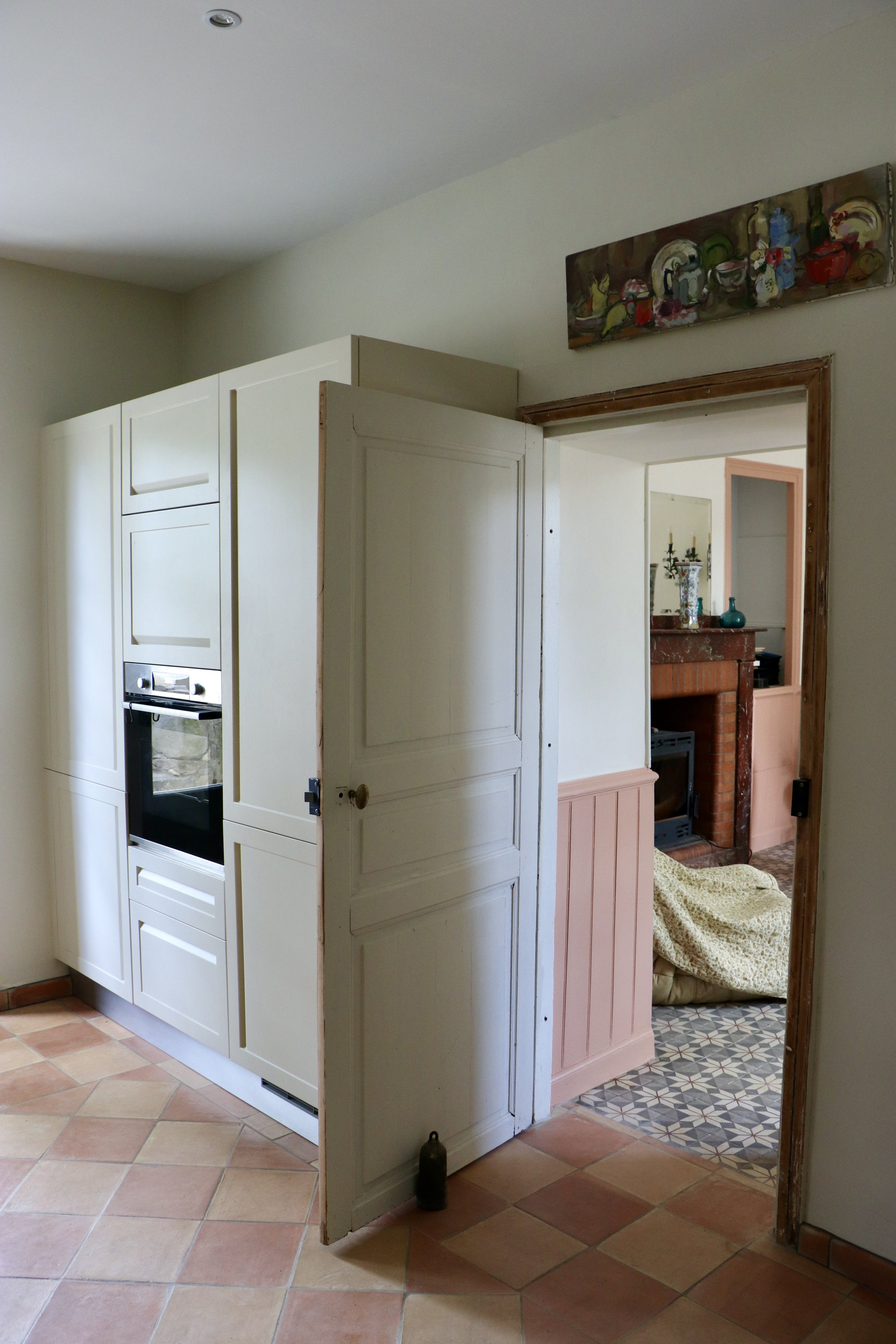 Vue intérieure d'une maison, avec une porte ouverte menant à une cheminée en pierre avec des candélabres et des décorations, canapé partiellement visible avec une couverture beige, mur blanc, sol en carreaux de terre cuite.