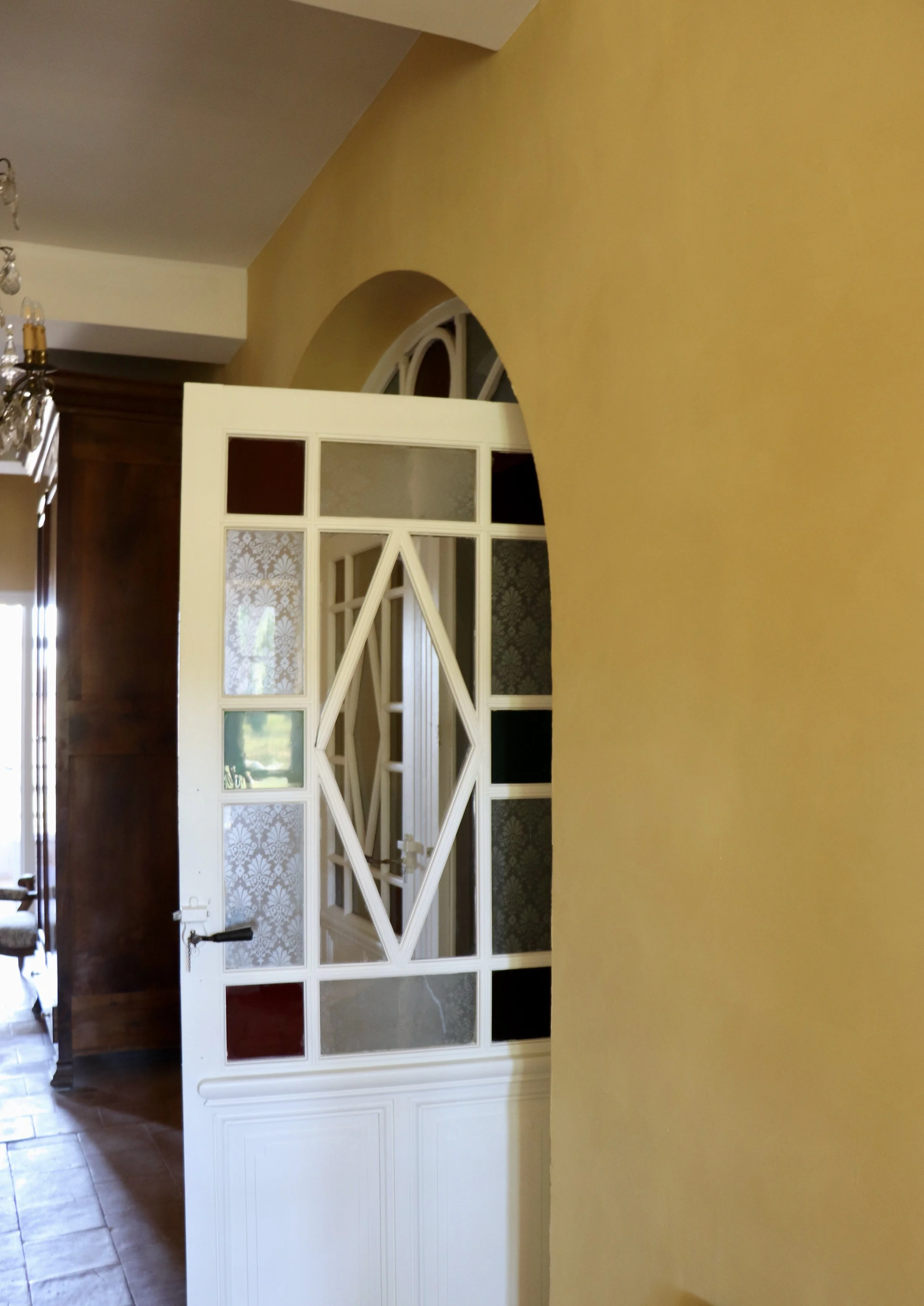 Une porte en bois blanc avec des fenêtres en verre coloré et en relief, dans un intérieur avec un mur jaune et un plafond blanc, laissant entrevoir une pièce avec du mobilier en bois.