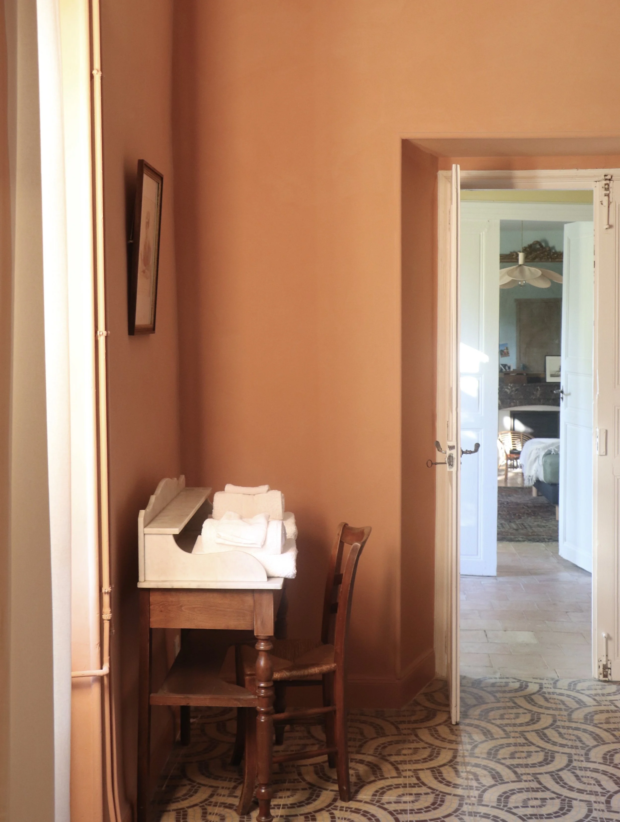 Intérieur d'une pièce avec un mur terracotta, un bureau en bois avec des serviettes blanches dessus, une chaise en bois, et une porte ouverte qui mène à une autre pièce avec un lit et un canapé visible. peinture à la chaux, carreaux de ciment
