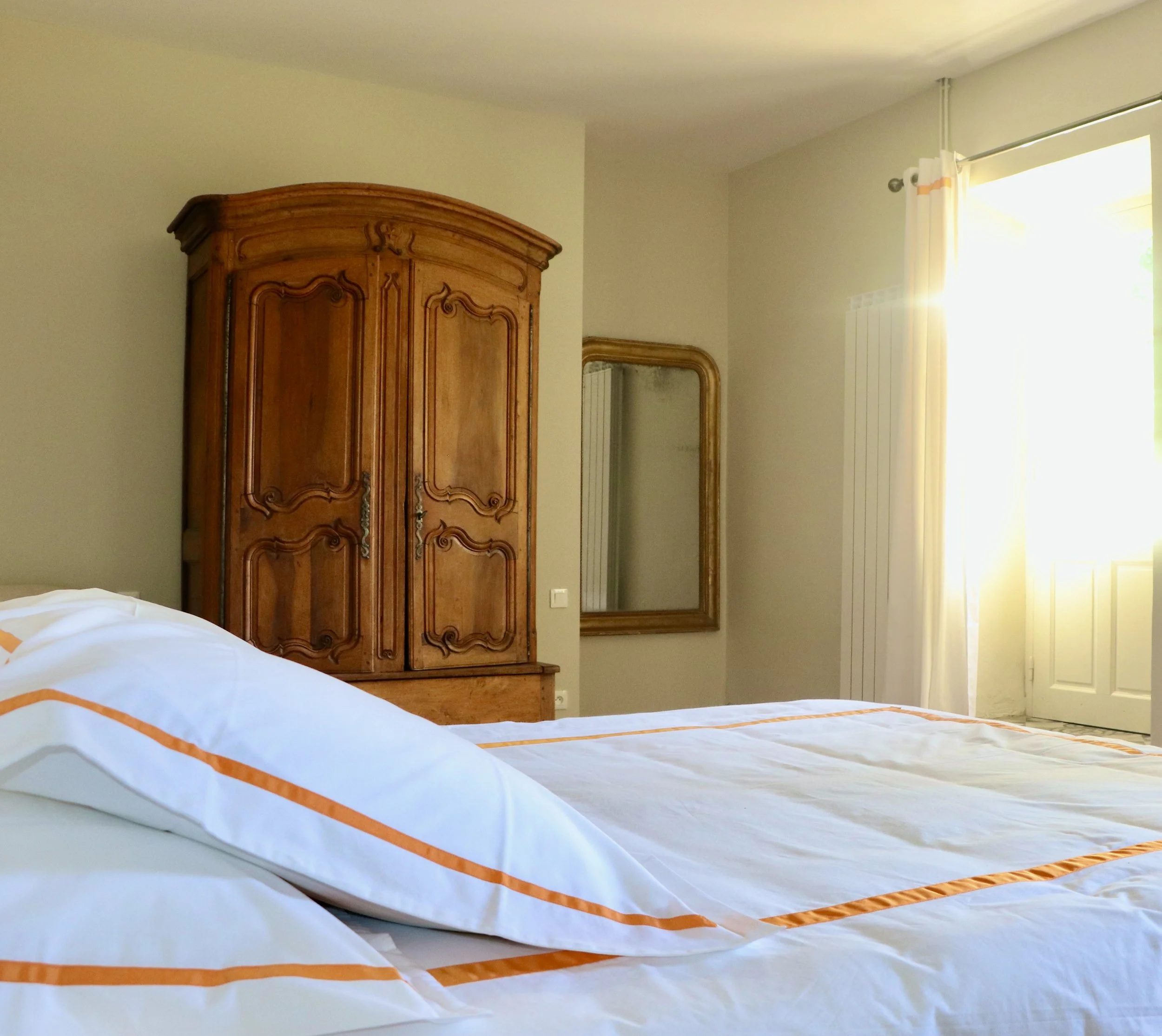 Chambre avec un lit en blanc et orange, armoire en bois, miroir, porte-fenêtre avec rideaux, lumière naturelle