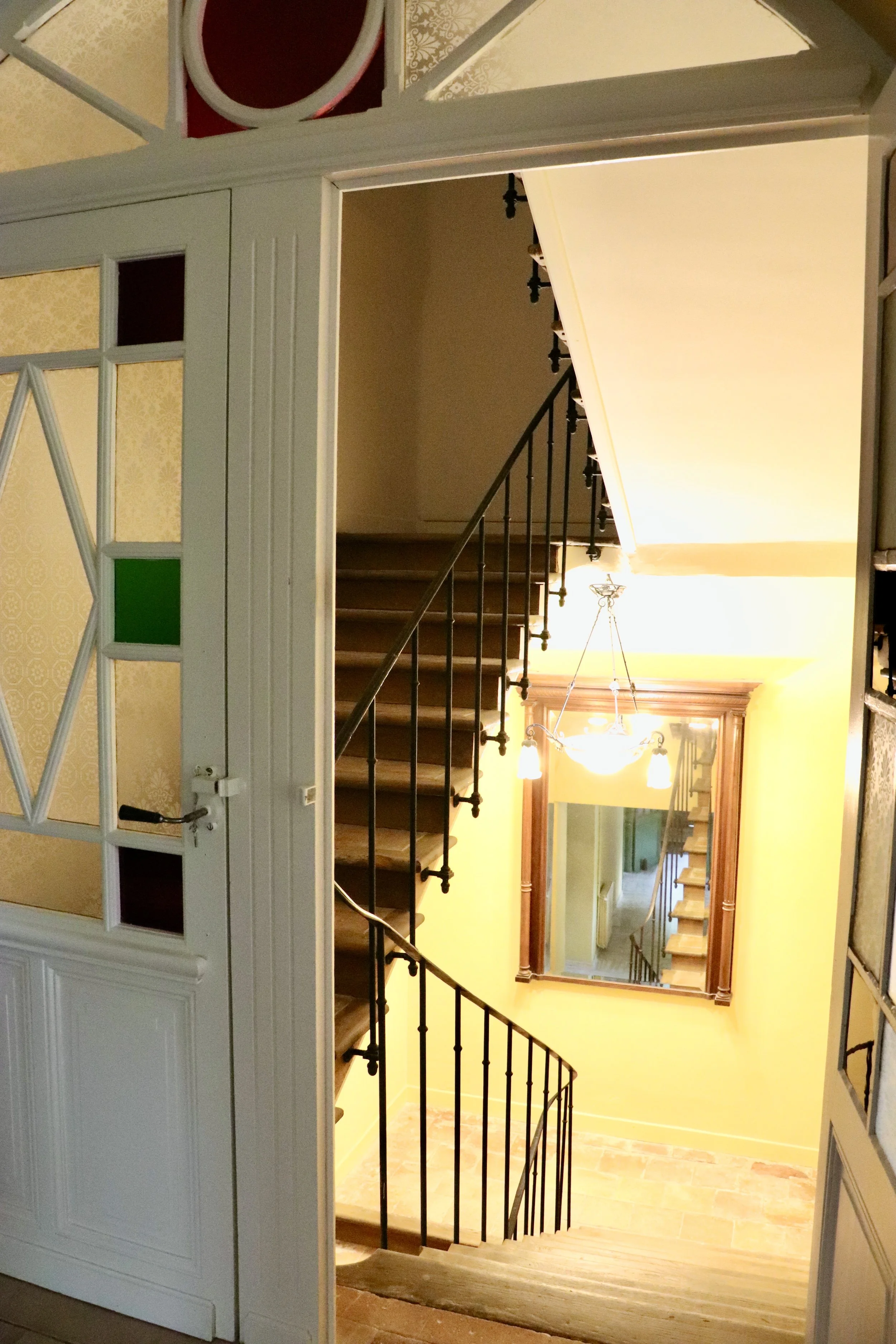 Vue intérieure d'un escalier en colimaçon avec une rampe en fer forgé, un miroir au mur, un chandelier suspendu et une porte avec de petites fenêtres colorées. verres colorés 1900