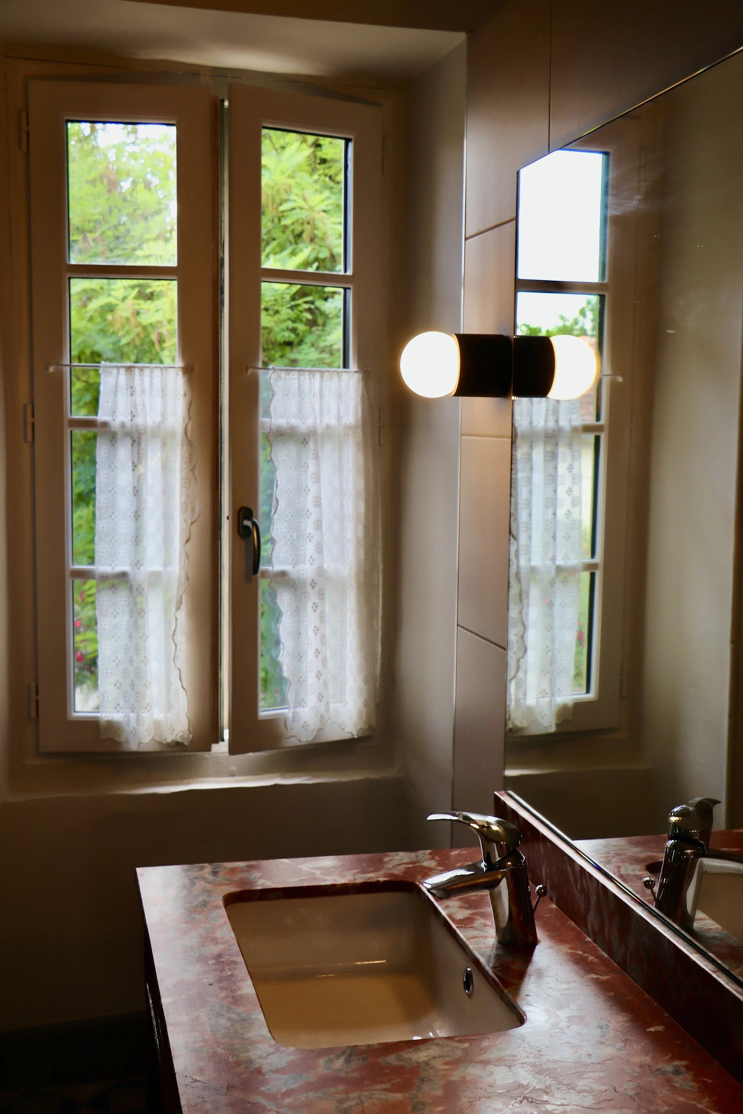 Lavabo avec robinet sur un comptoir en marbre rouge, miroir et fenêtre avec rideaux en dentelle blanche, vue de l'intérieur d'une salle de bain. Marbre de Caunes