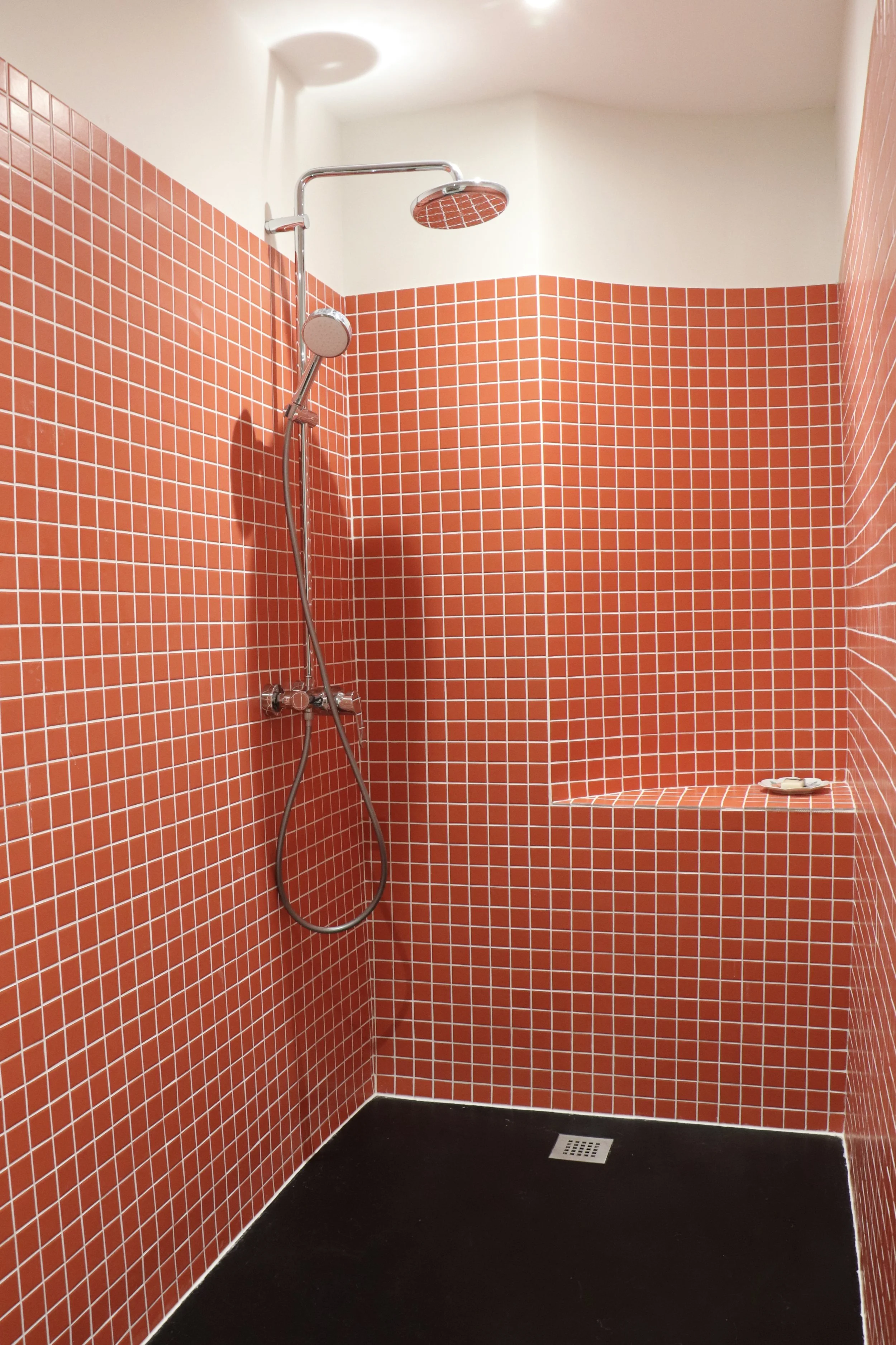 Une douche avec carreaux rouges, une pomme de douche au plafond, une étagère intégrée avec quelques objets. carreau mosaique douche