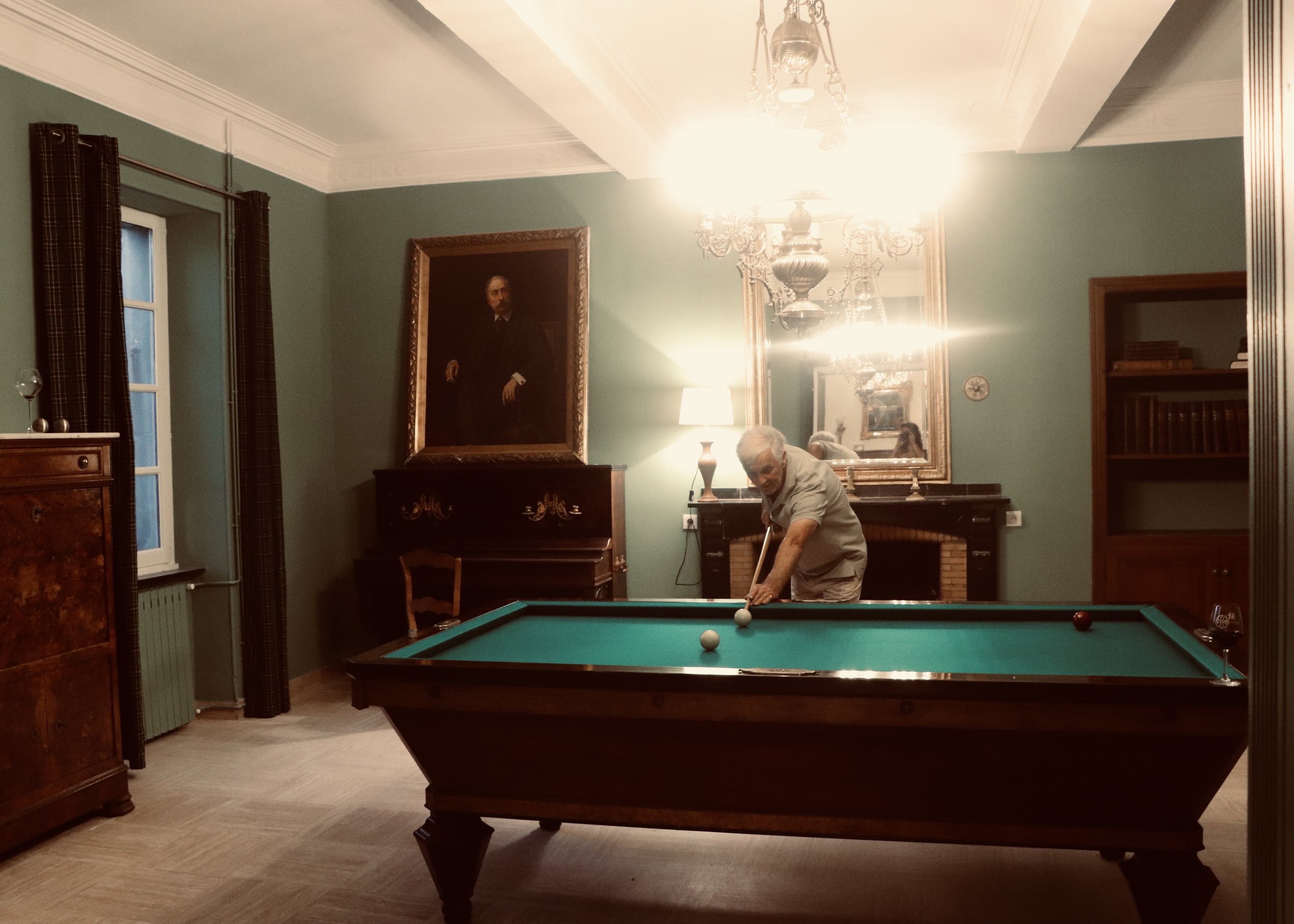 Un homme âgé joue au billard dans une salle de style classique, avec un grand portrait encadré et un miroir, une cheminée, un lampadaire, et des meubles en bois. Billard Français