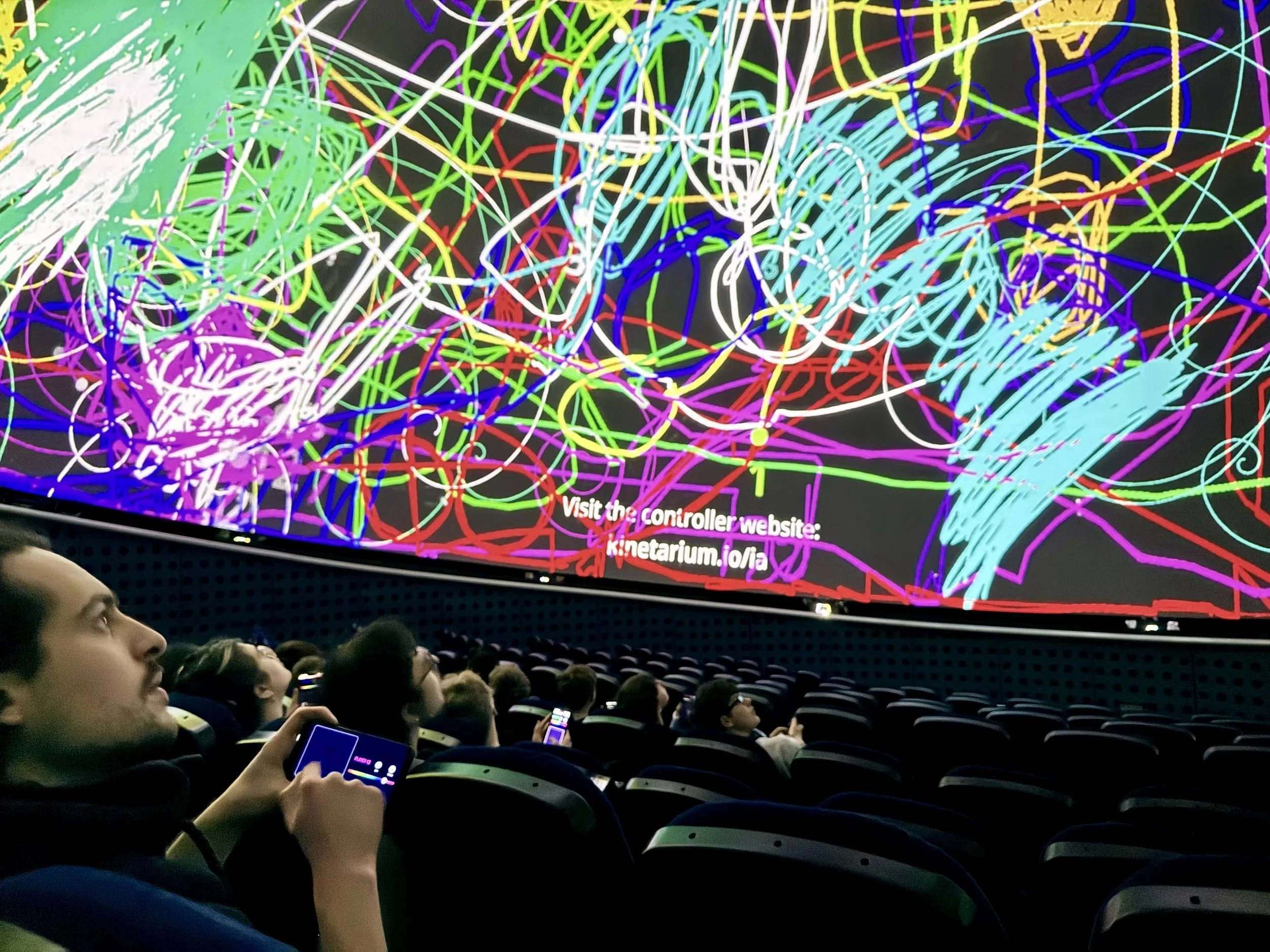 Besuchende in einem Planetarium spielen das Kinetarium Game Kuppelgraffiti und erzeugen über ihren Smartphones gemeinsam ein buntes Bild mit einer Vielzahl von leuchtenden, wirbelnden Linien in verschiedenen Farben.