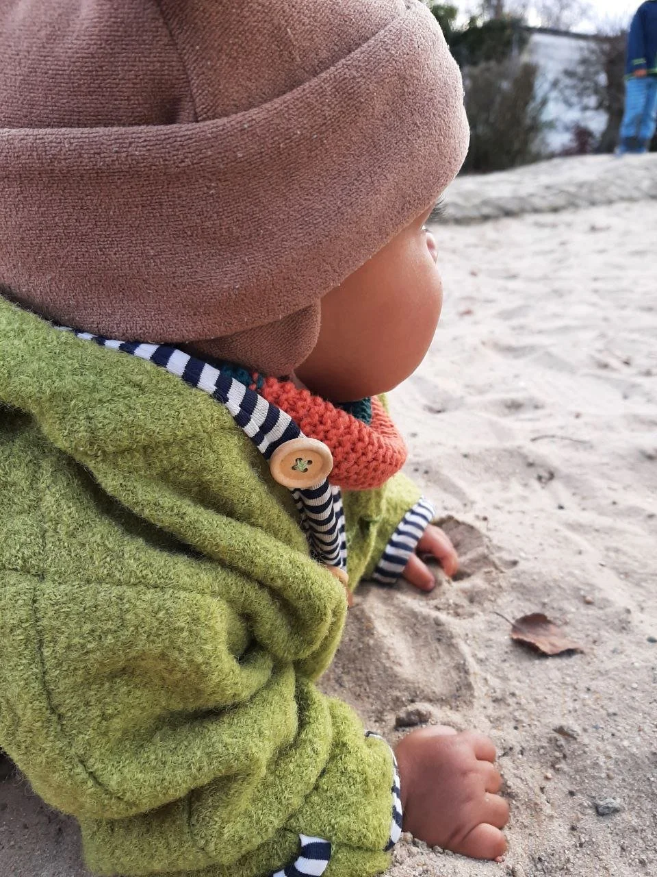 Kleines Kind, das im Sand spielt. Es hat einen grünen Anzug und eine braune Mütze an. Im Hintergrund sind Pflanzen zu erkennen.