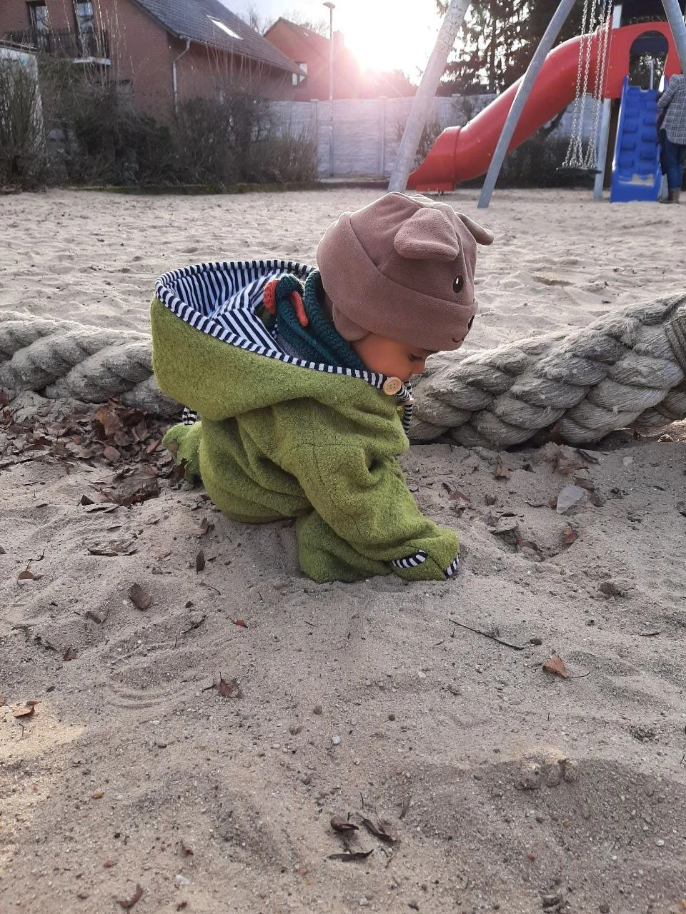 Kleines Kind in einem grünen Mantel und einer braunen Mütze mit Tierohren spielt im Sand auf einem Spielplatz, im Hintergrund ein Rutschenturm.
