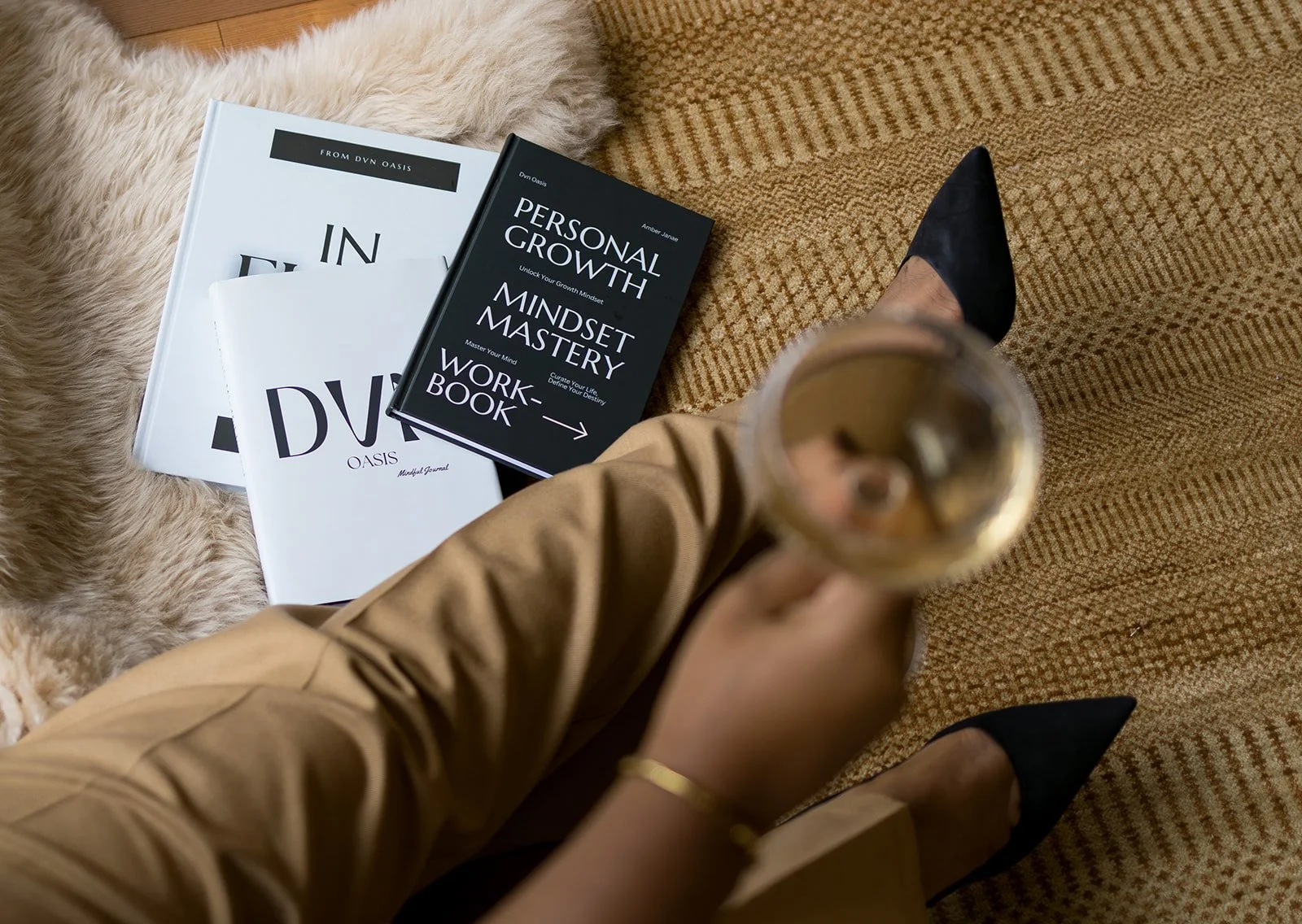 A person wearing black pointed-toe shoes and tan pants sitting on a carpet, holding a glass of beverage. Nearby, on a fur rug, are three books about personal growth, mindset, and workbooks.