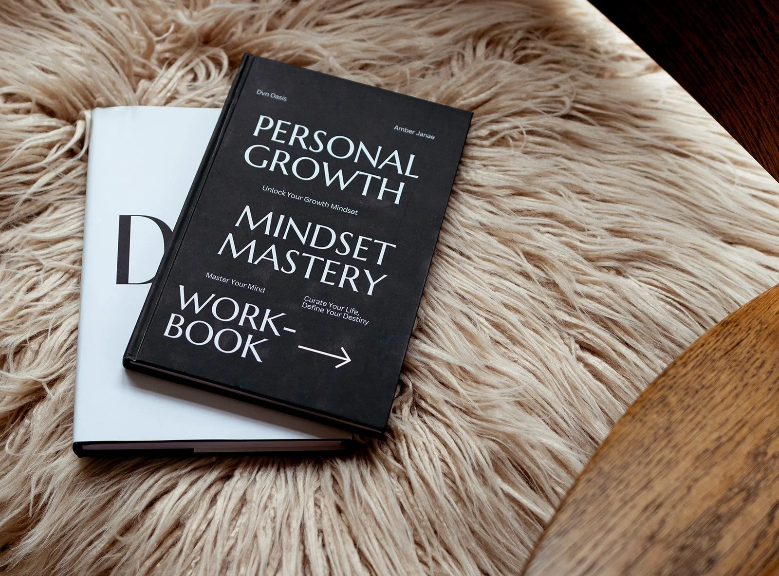A black book titled 'Personal Growth Mindset Mastery' and a white notebook resting on a fluffy beige rug, with a wooden table corner visible on the right.
