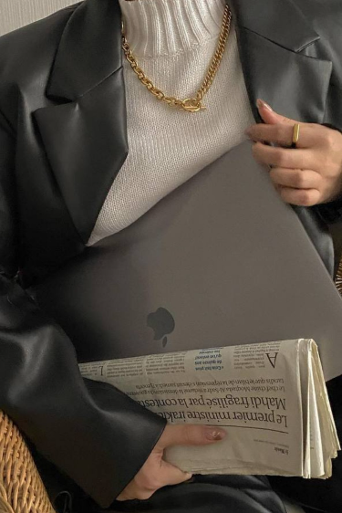 Person holding a laptop and a newspaper, wearing a black blazer and a gold chain necklace.