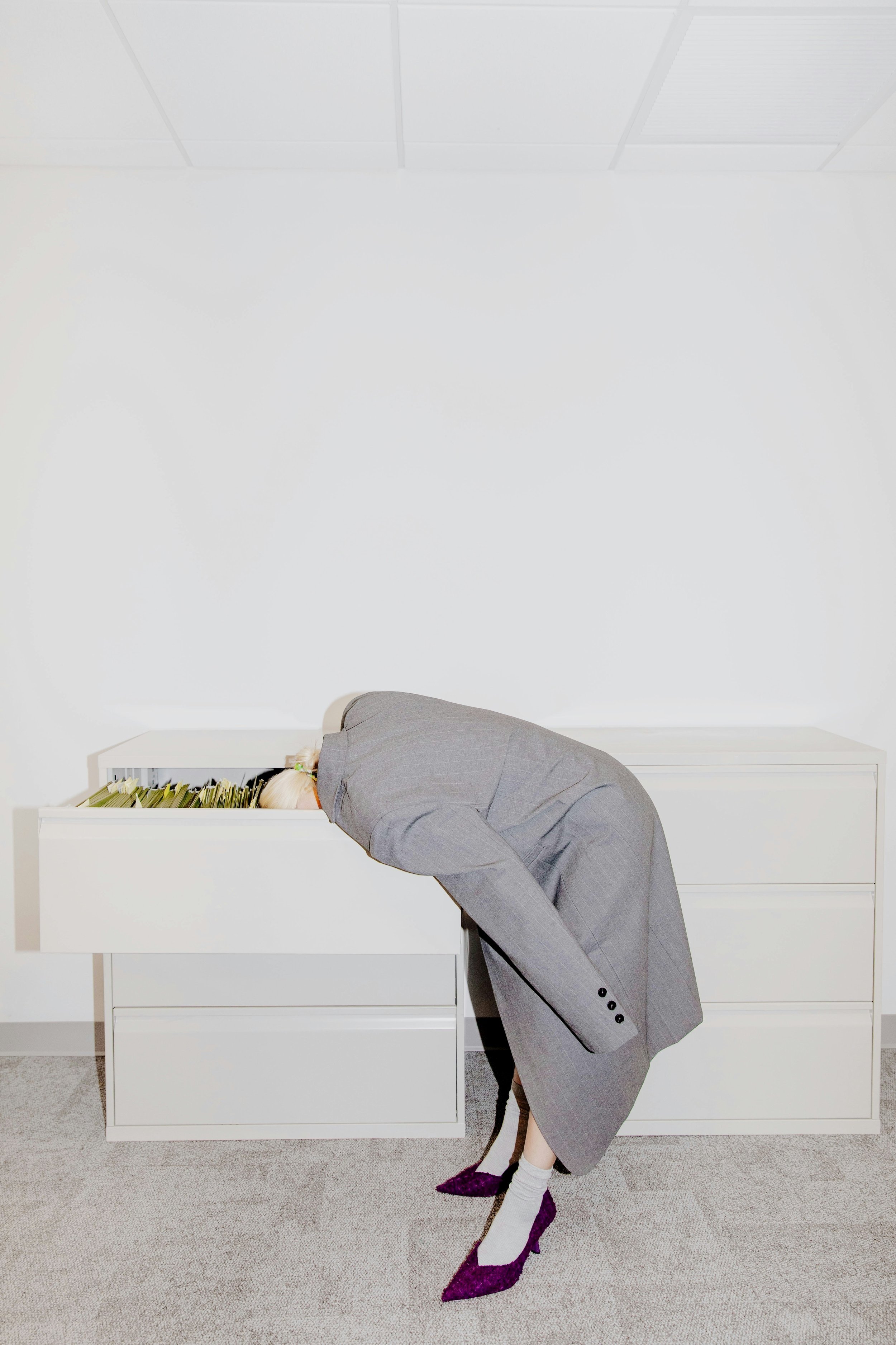 Person in a gray suit leaning into a filing cabinet with head inside, wearing purple high heels and white socks.