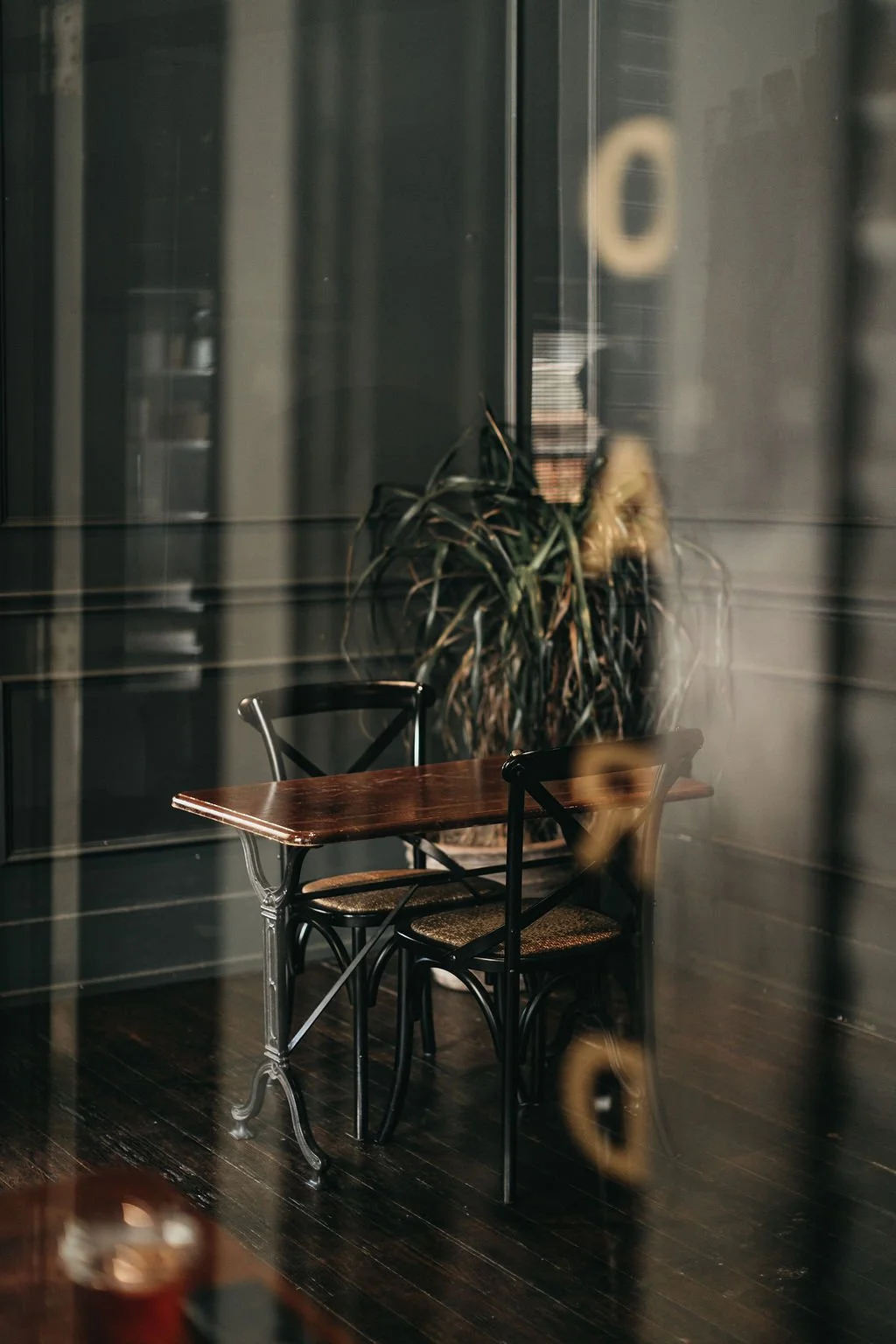 A small wooden table with two black chairs, and a large leafy plant behind it, viewed through a glass window with reflections in a cozy indoor setting.
